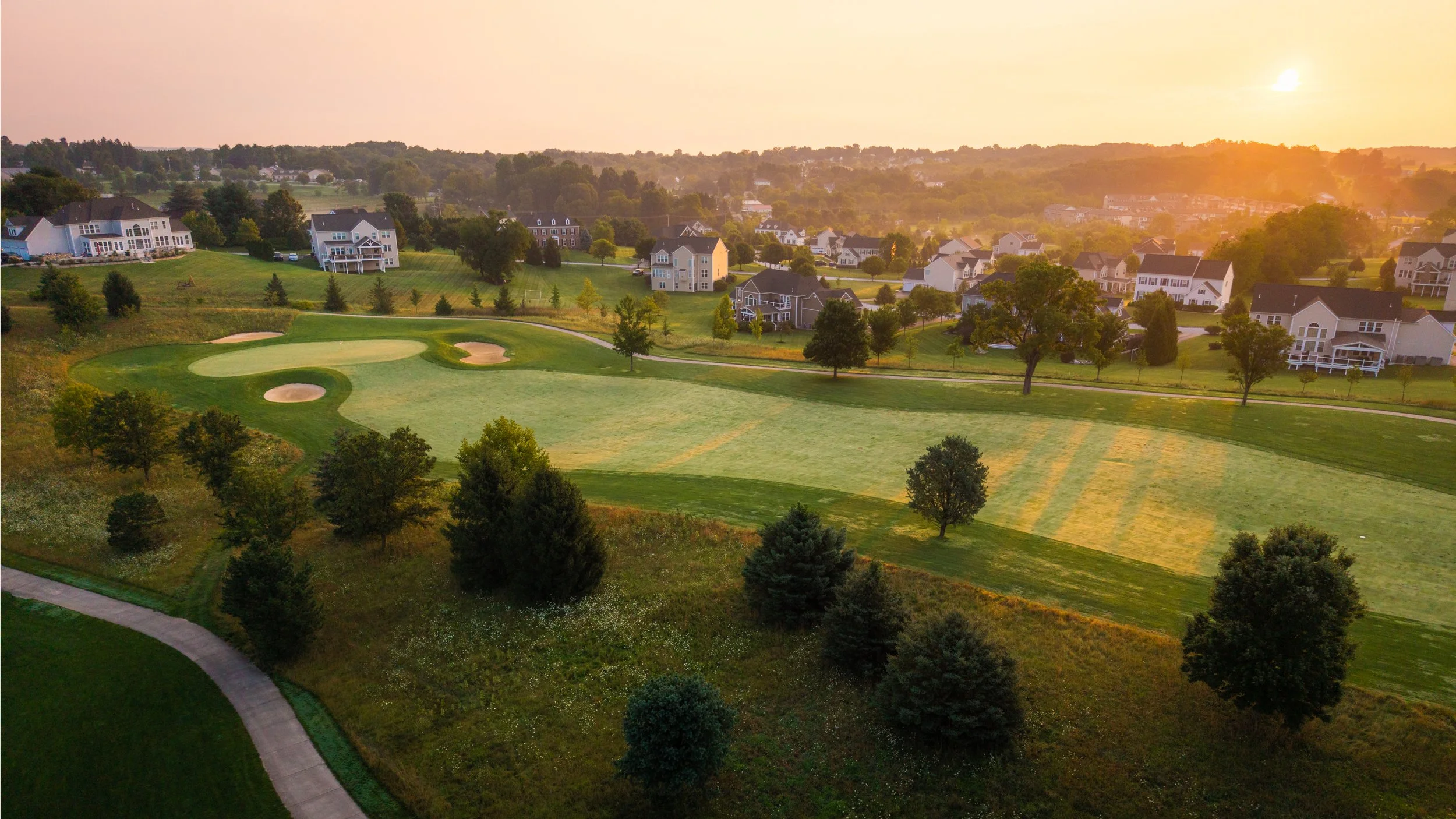 A scenic aerial view of a golf course with green fairways, sand traps, and surrounding trees, adjacent to residential houses at sunset.
