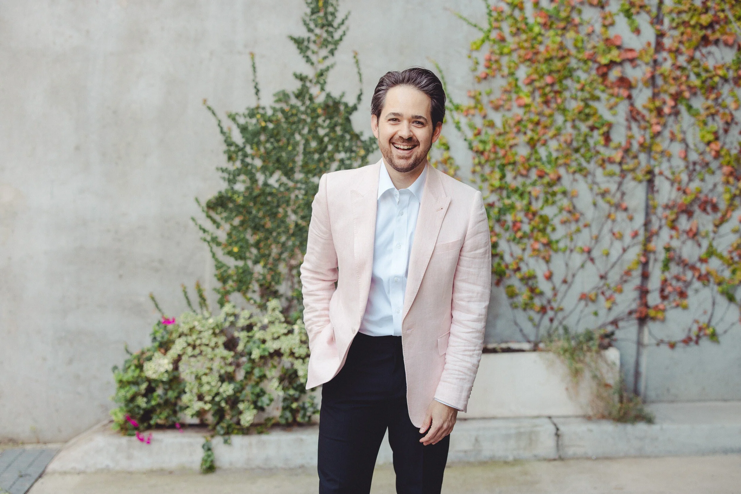 A man smiling and wearing a pale pink blazer with a white shirt, standing outdoors in front of a gray wall with green and reddish leaves climbing it, and a small flower arrangement in front of him.