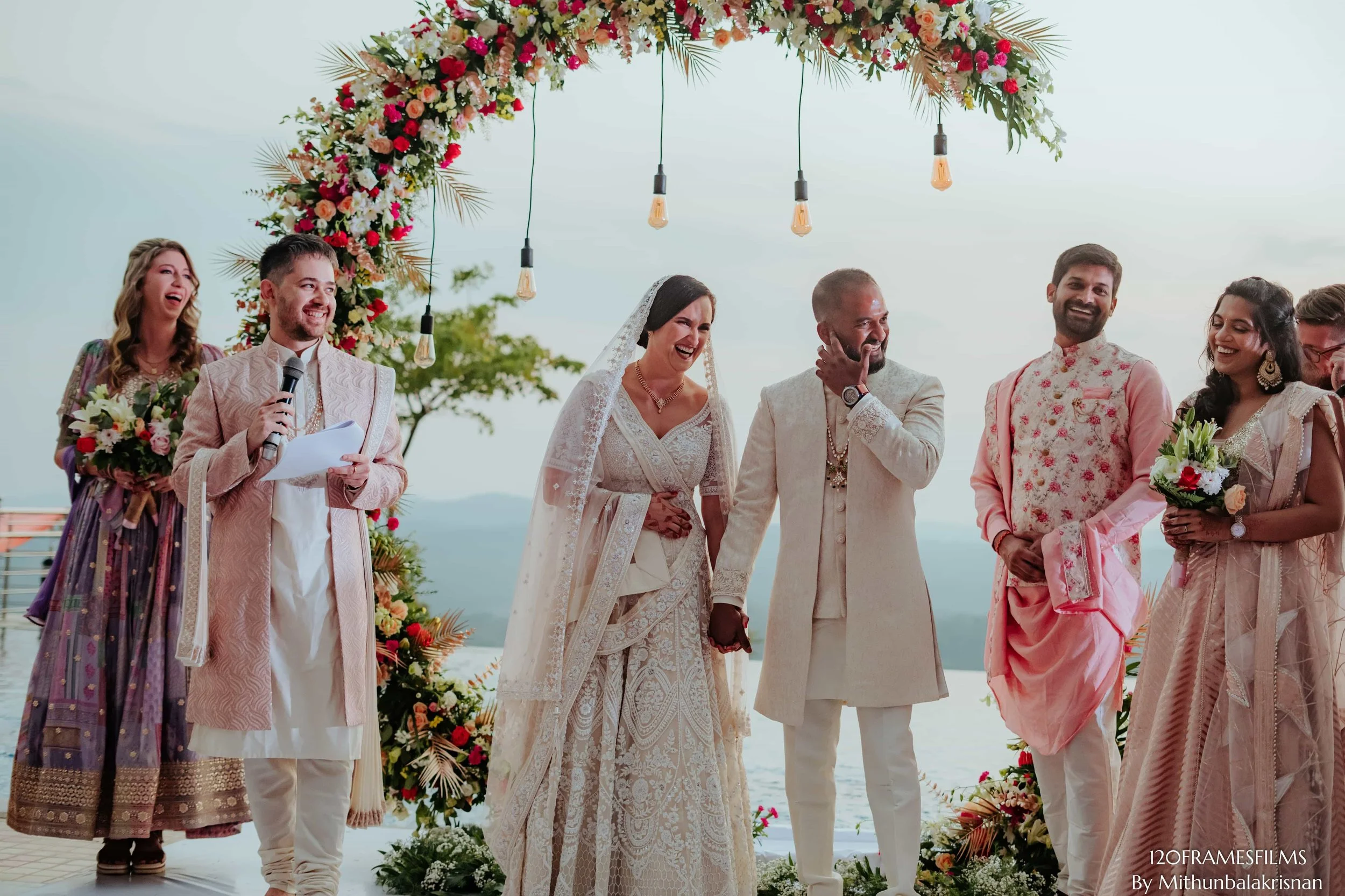 A wedding ceremony taking place outdoors with the couple holding hands under a floral arch, surrounded by friends and family, dressed in traditional Indian attire, with a lake or ocean in the background.