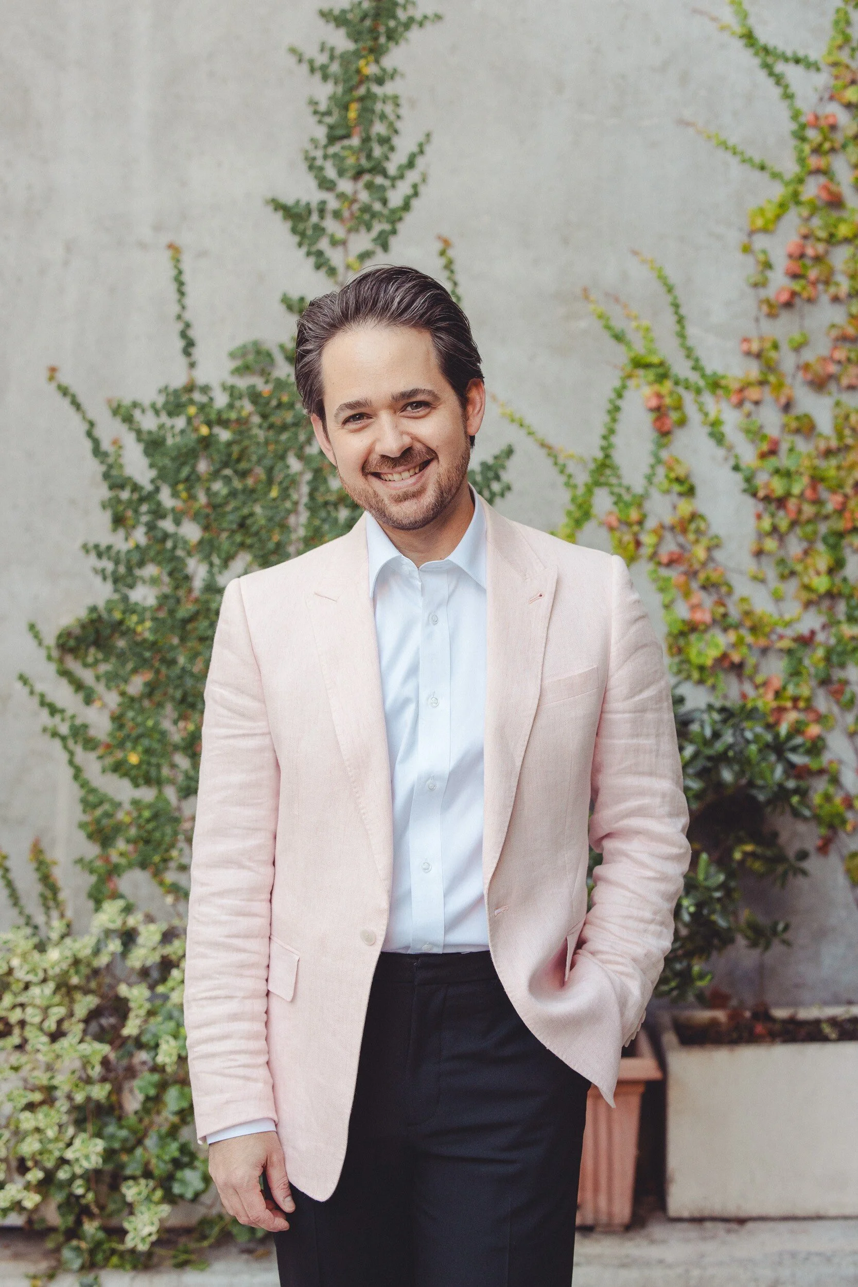 A man in a light pink blazer, white shirt, and black pants smiling and standing outdoors in front of a concrete wall with green and reddish vine plants.