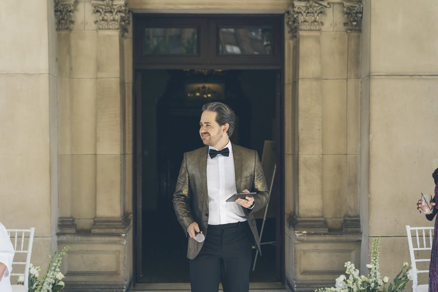 A man dressed in a tuxedo with a gold blazer holding a microphone and a mobile phone, standing in front of a building entrance.