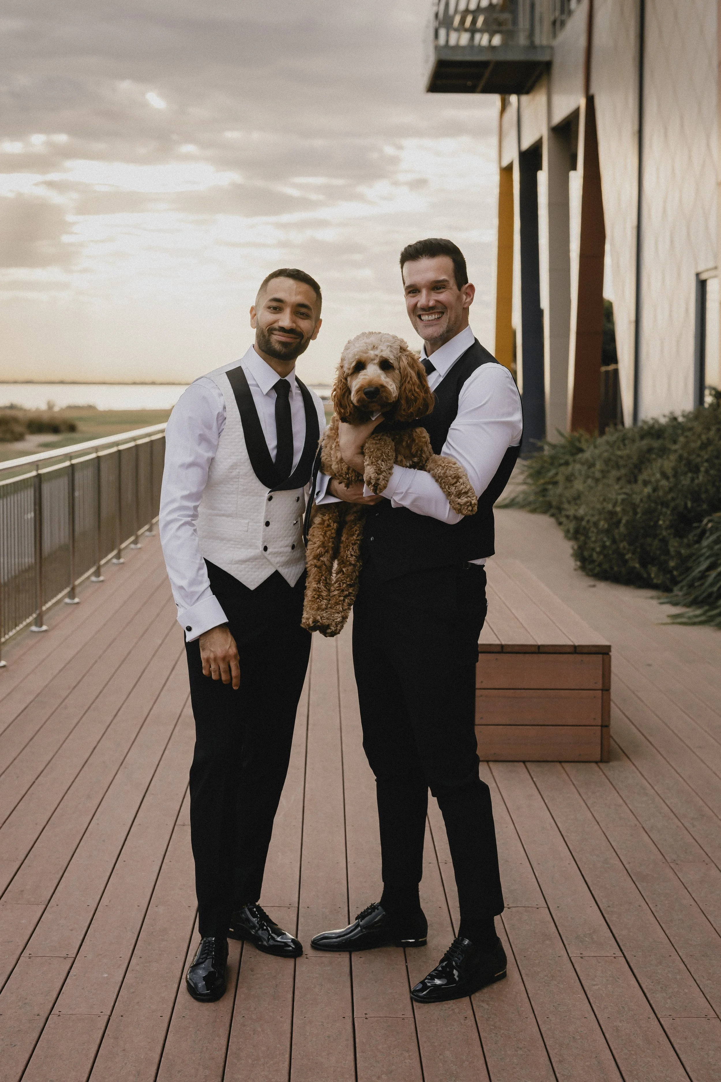 Two men in tuxedos standing on a wooden deck, holding a brown dog with curly fur, during sunset with a riverview in the background.