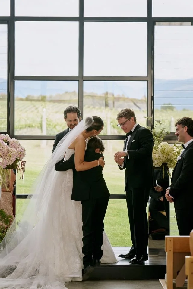 Three men in suits, a bride in a white dress hugging a junior groomsman who is handing over the rings