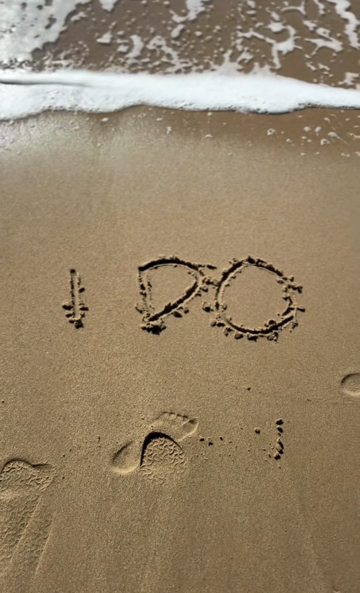 The image shows the words 'I ❤️ P' written in the sand at the beach with footprints nearby and a wave approaching the shore.