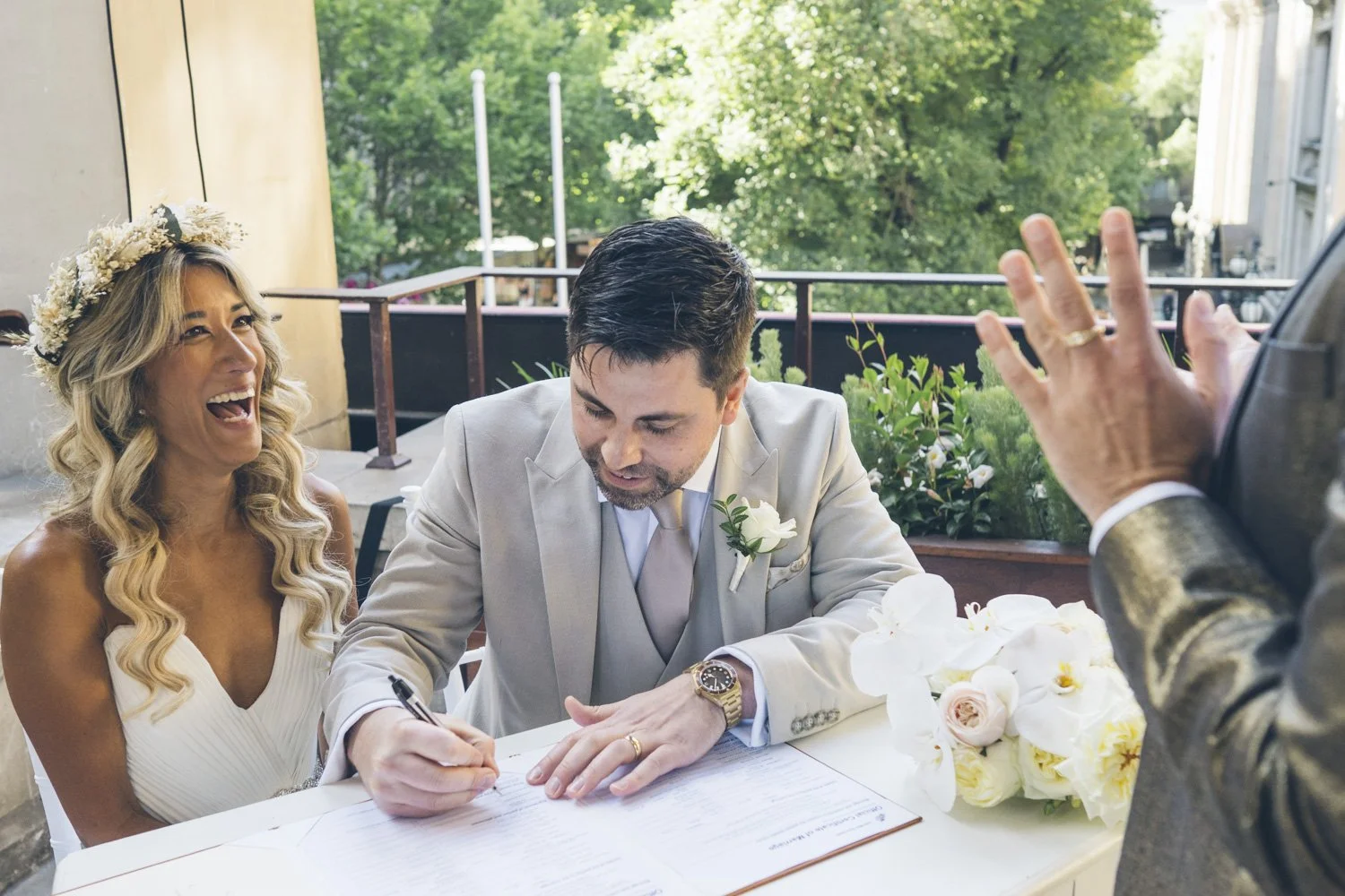 A couple signs a marriage document during their wedding ceremony, with a woman laughing and an officiant raising a hand nearby. The bride has long blonde hair with a flower crown, and the groom has dark hair, wearing a light-colored suit with a white boutonniere. A bouquet with white flowers is on the table, outdoors with greenery in the background.