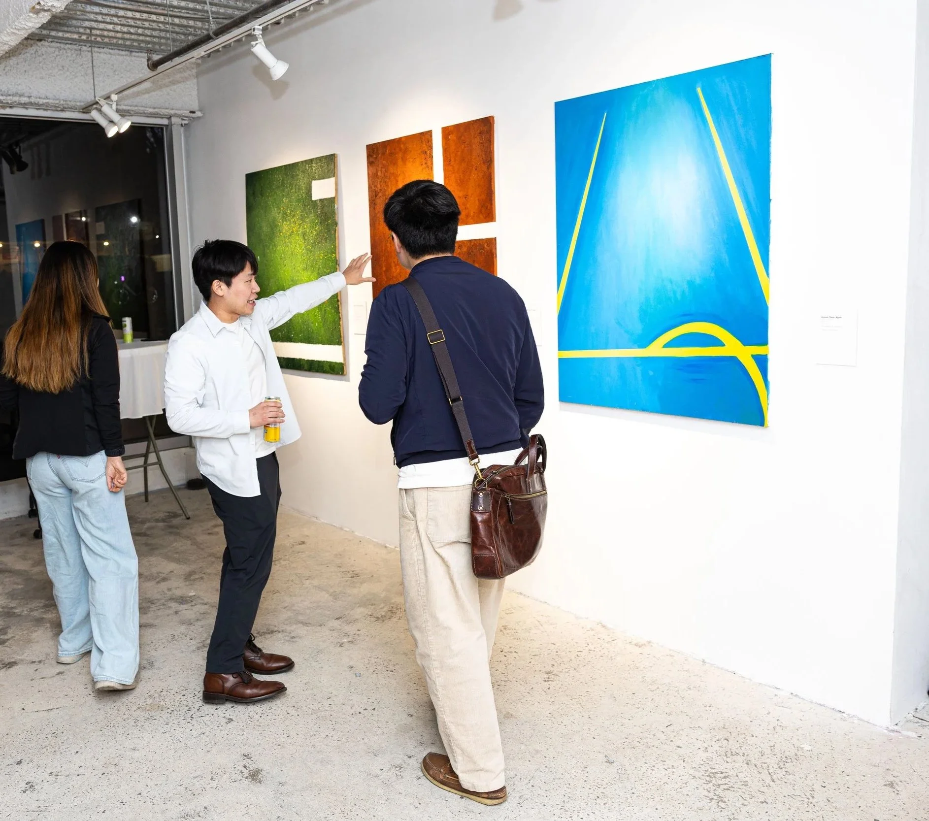 Three people, two men and one woman, are talking in an art gallery. The woman has long hair and is wearing a black jacket and blue jeans. One man has short black hair, wearing a white shirt, and holding a yellow can. The other man has black hair, wearing a navy jacket and beige pants, with a brown bag over his shoulder. They are standing near colorful abstract paintings on a white wall. The gallery has a concrete floor and visible ceiling pipes.