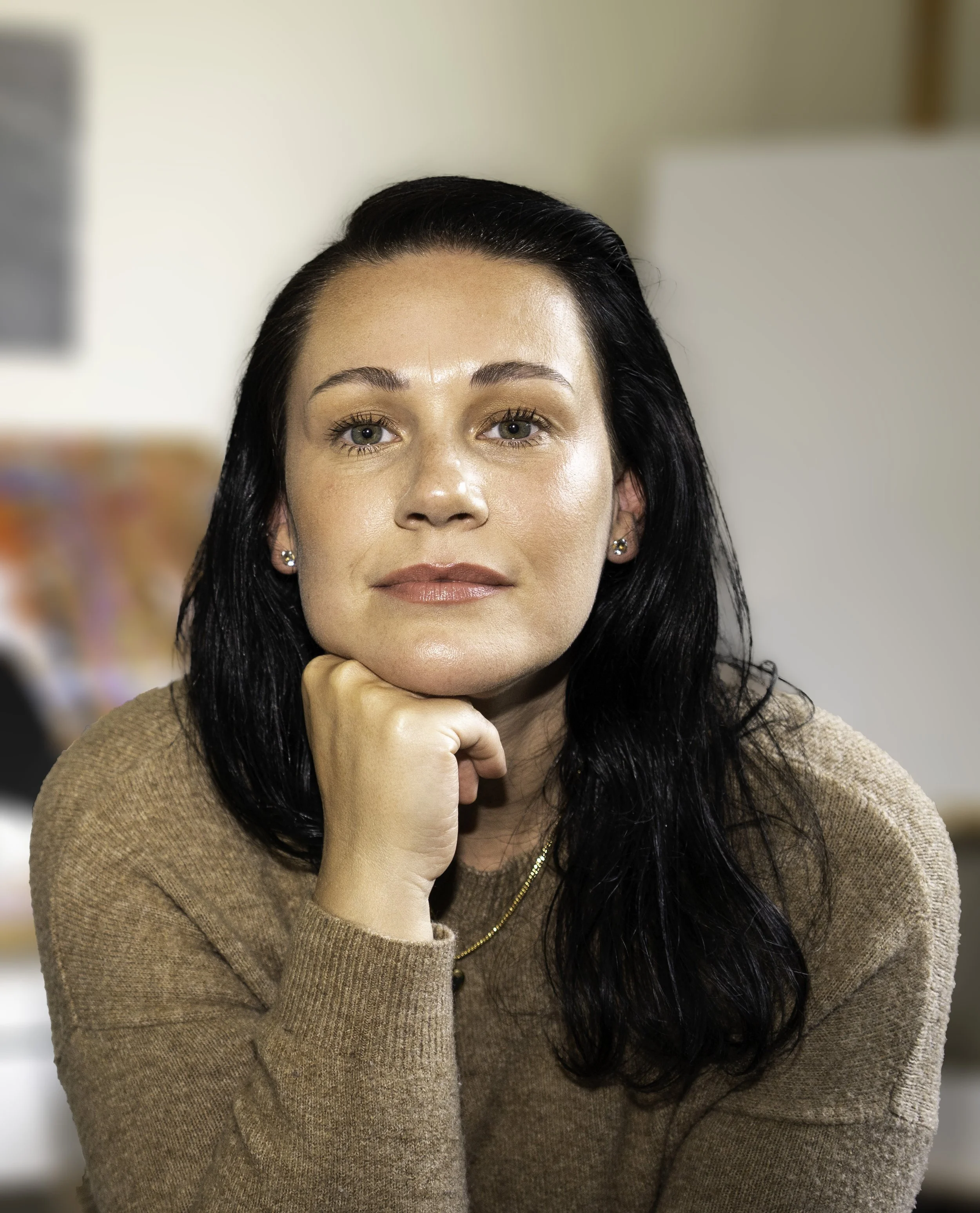 A woman with dark hair, light skin, and green eyes, resting her chin on her hand and wearing a beige sweater, in a softly blurred indoor setting.
