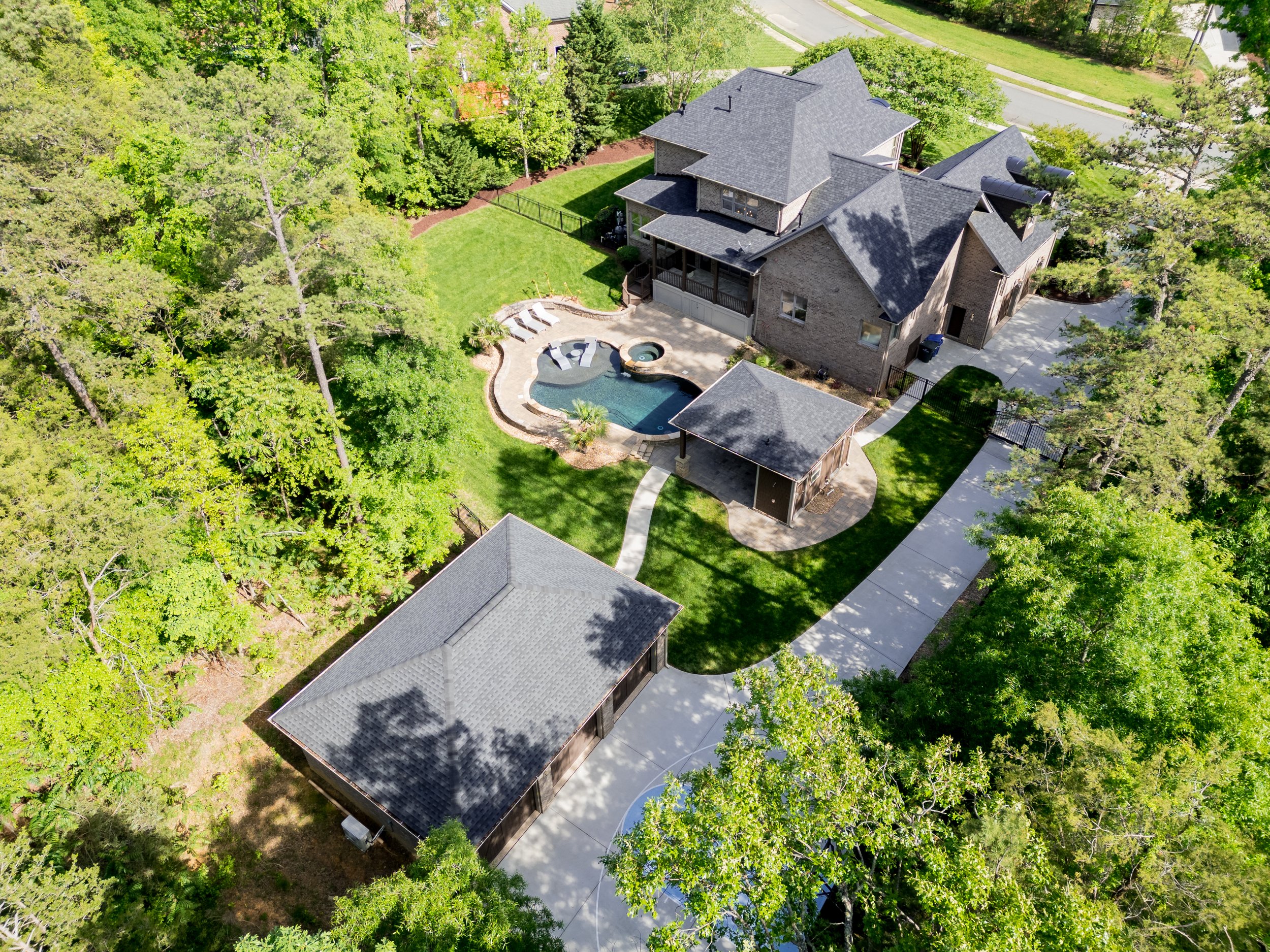 Aerial view of a large house with a dark roof, a swimming pool with a curved design, a detached garage, and surrounding trees