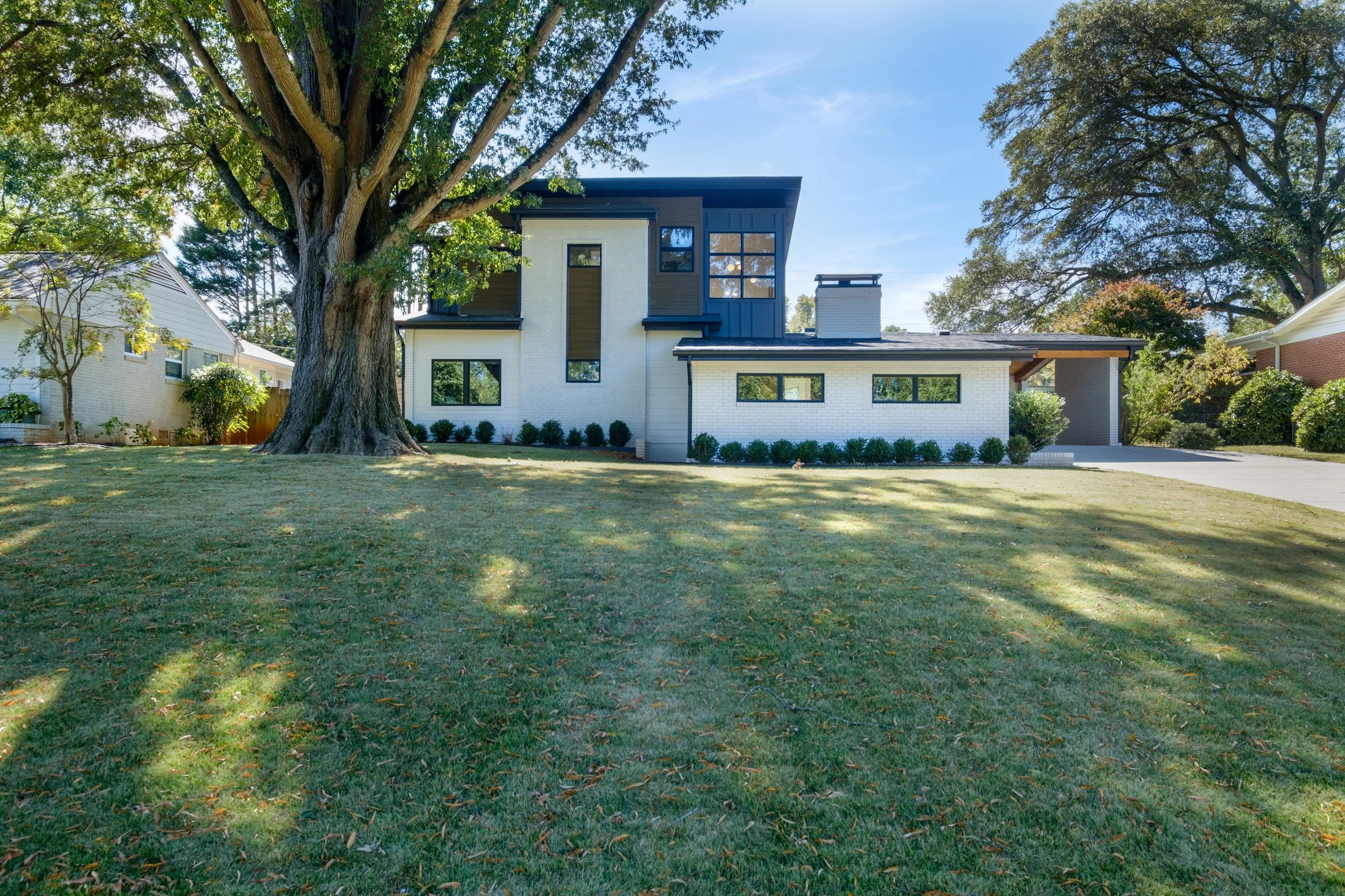 Modern two-story white and dark blue house with large windows, covered carport, and a large tree in the front yard