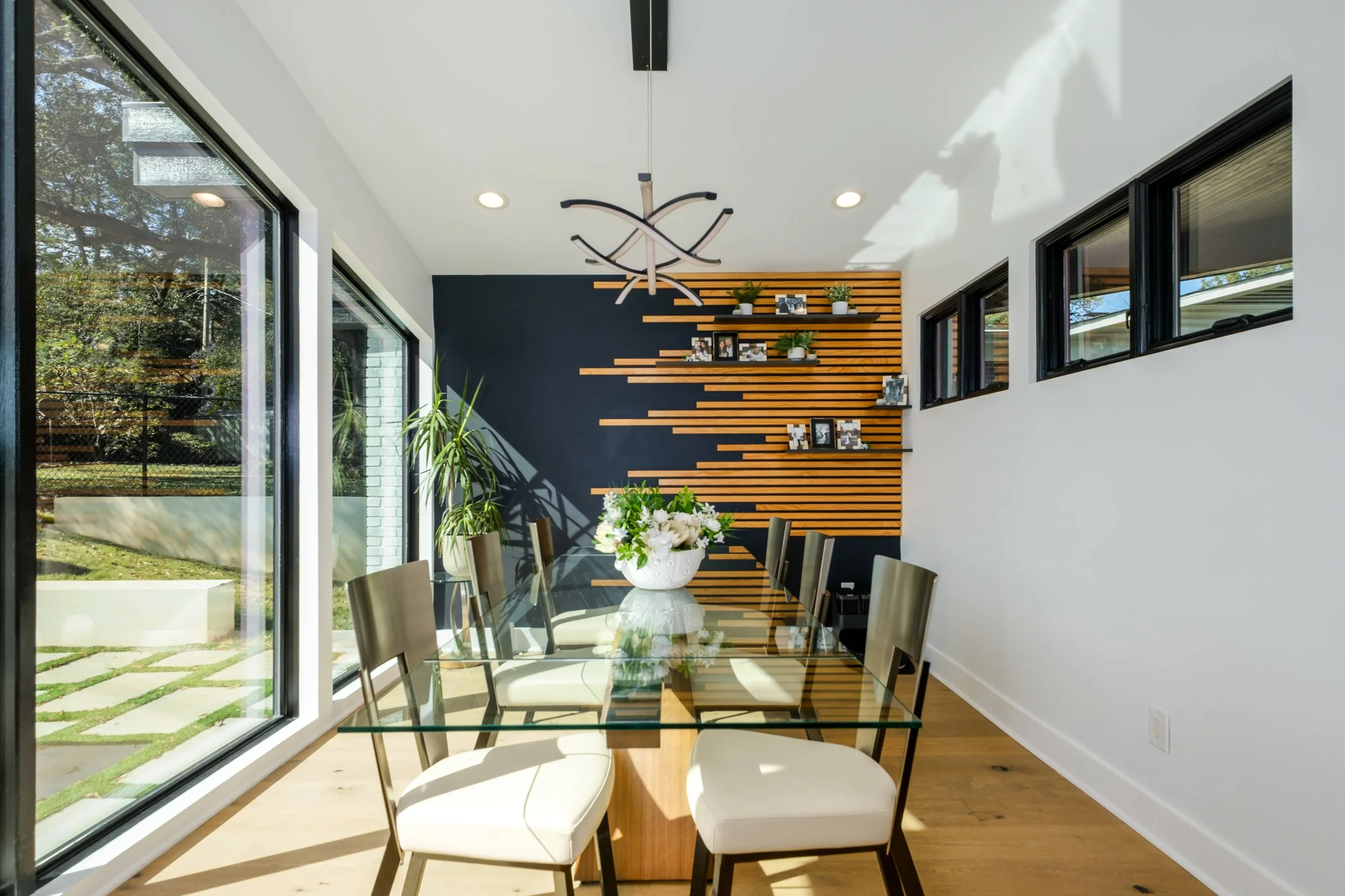 Modern dining room with a glass table, neutral chairs, a dark blue accent wall with horizontal wood slats, and large windows