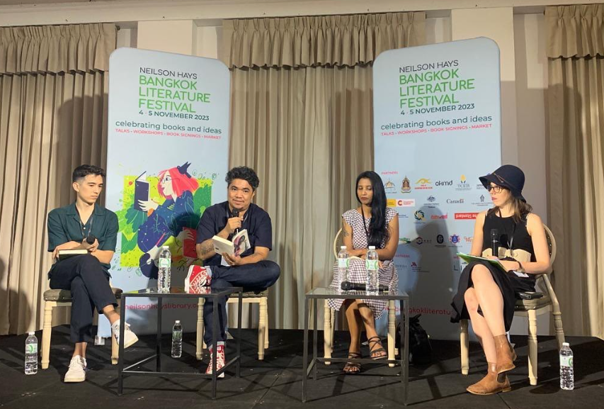 Panel discussion at the Bangkok Literature Festival, featuring three authors and Lisa Martin seated on stage with microphones and books, with banners in the background displaying event details and logos.