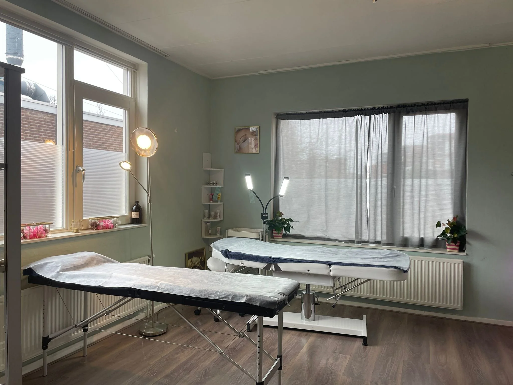 A massage therapy room with two massage tables, one with disposable sheet, in a room with large windows, curtains, potted plants, a standing lamp, and soft lighting.