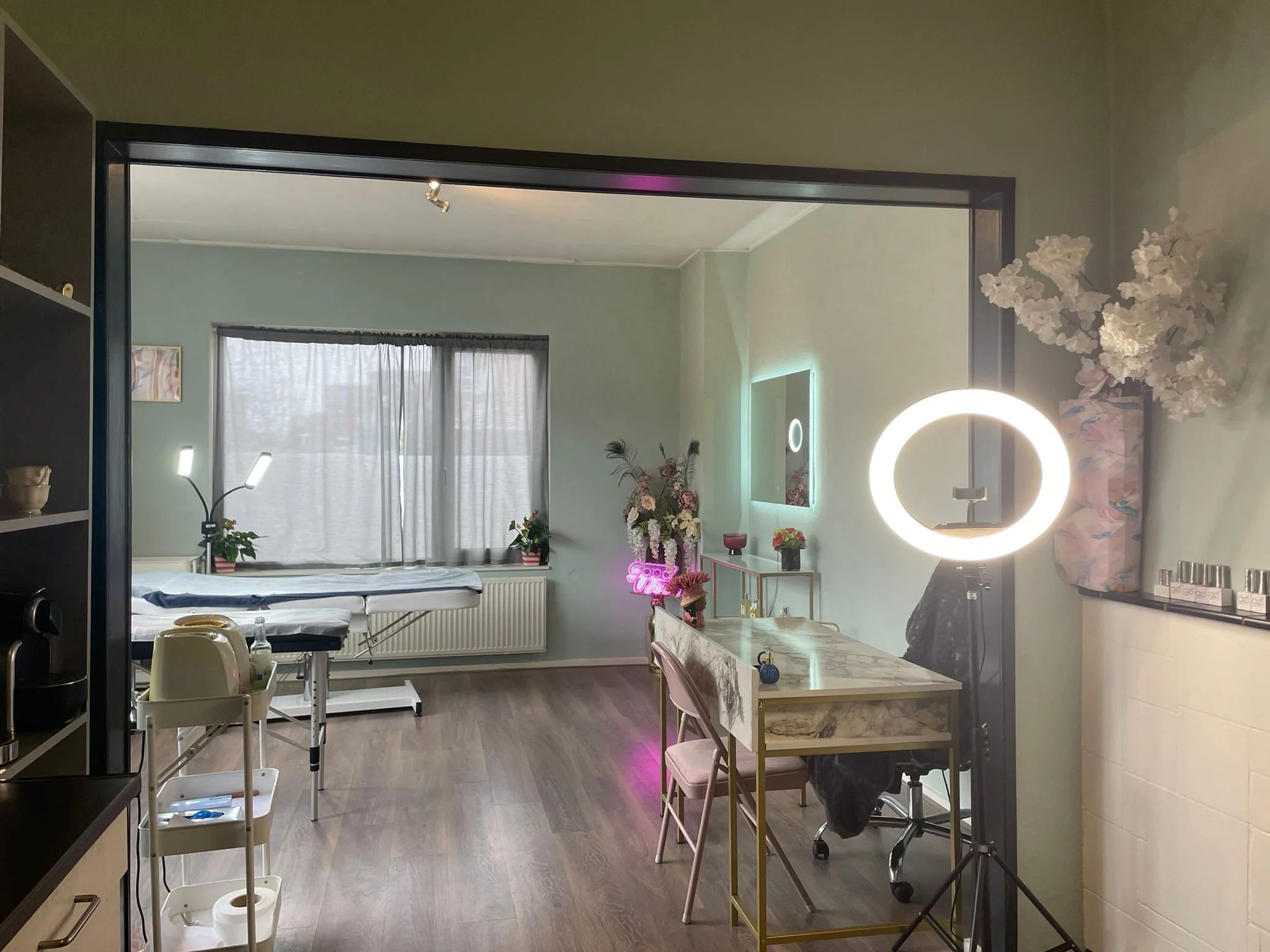 Interior of a room with a bed by the window, a mirror with LED lighting, a ring light, a marble desk with a pink chair, and decorative flowers.