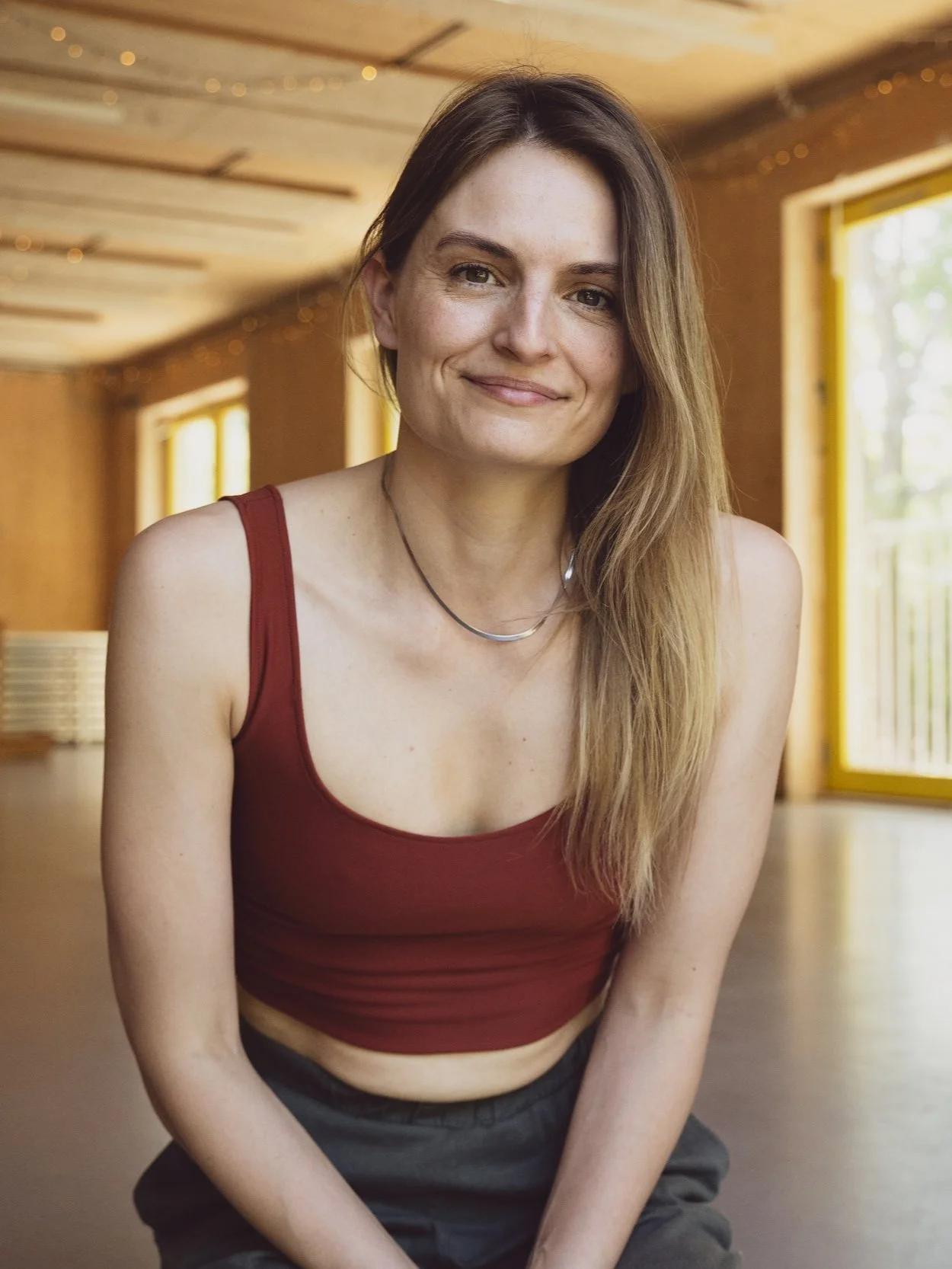 A smiling woman with long hair wearing a red tank top and dark pants in a wooden room with large windows.