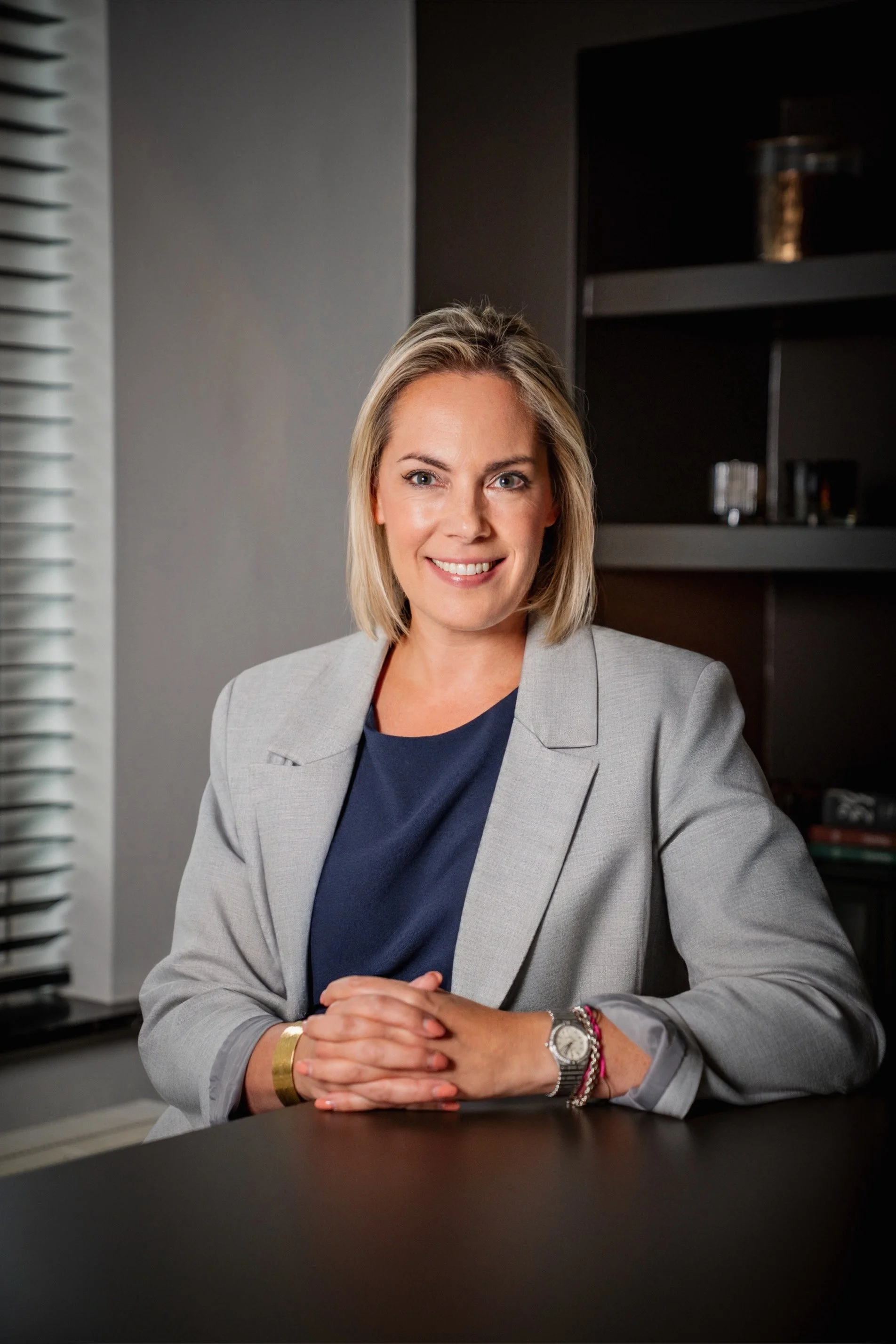 Een vrouw met schouderlange blonde haar, gekleed in een witte blouse met een strik, staat met gekruiste armen in een hal met hoge gewelfde plafonds en klassieke architektonische details.