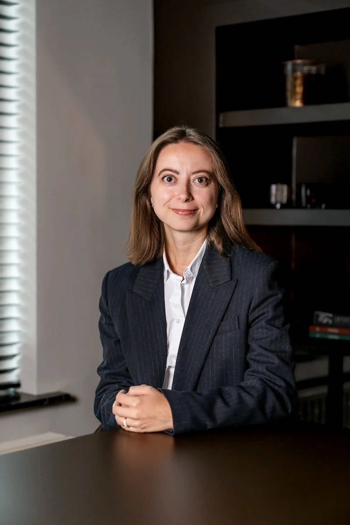 Een zakelijke vrouw met gekruiste armen staat in een elegante binnenruimte, gekleed in een zwart colbert en witte blouse.