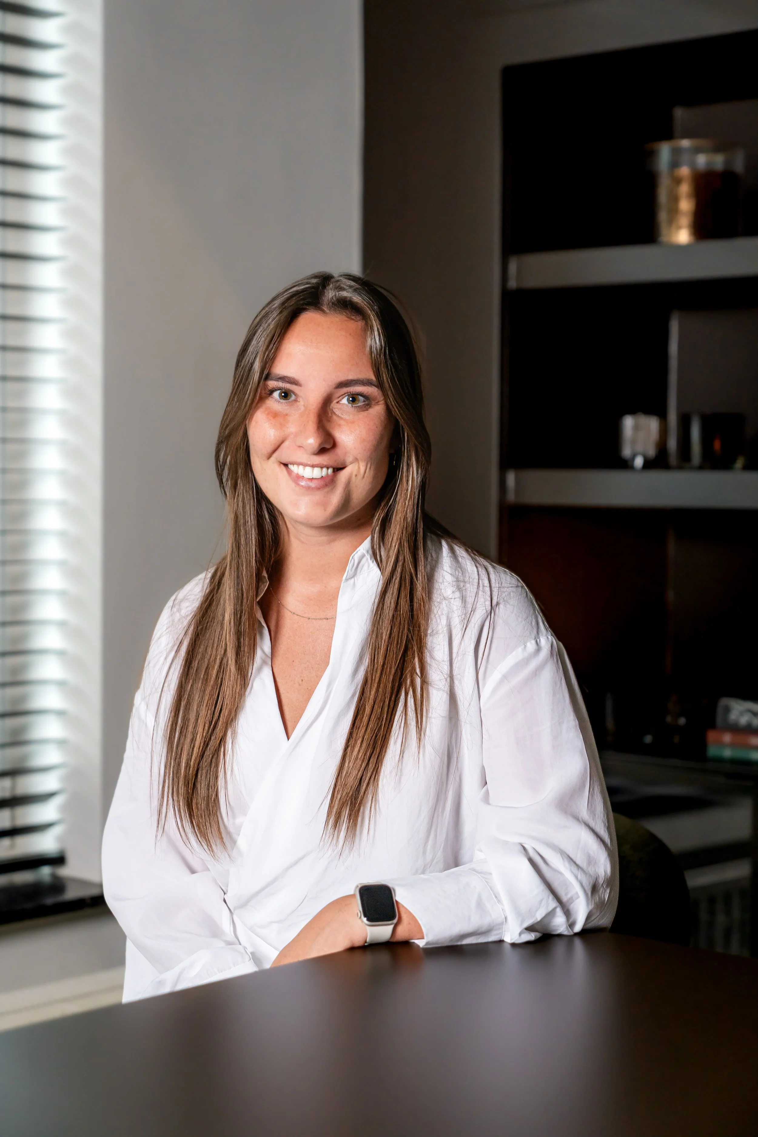 Portret van een jonge vrouw met lang, recht bruin haar, draag een witte blouse met een textuur en zwarte broek, staand in een elegante, warme interieurruimte.