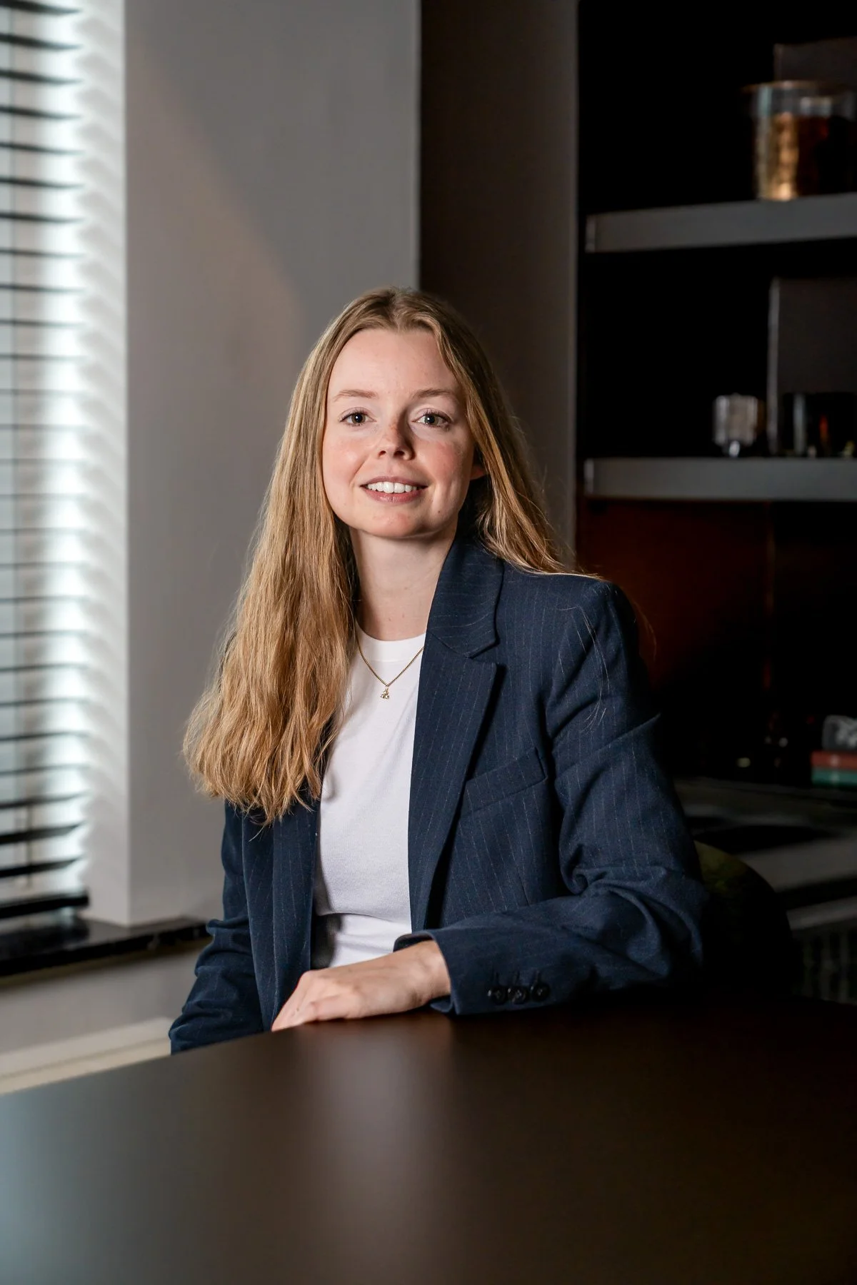 Een vrouw met lang, golvend bruin haar, glimlachend, staand in een elegante, sfeervolle kamer met houten vloeren en een boogdeur op de achtergrond, gekleed in een wit satijnen blouse en zwarte broek.