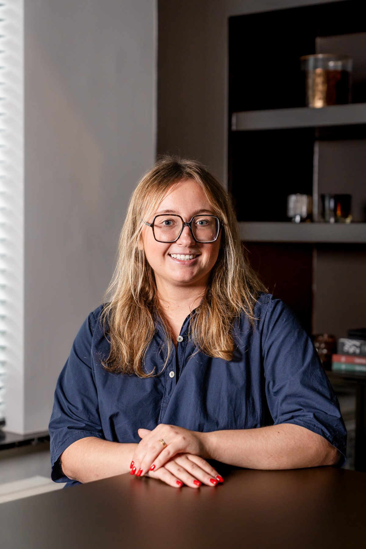 Portret van een jonge vrouw met bruin haar en brillen, gekleed in een zwarte trui met kleurrijke bloempatronen, standing in een interieur met houten details en zwarte stoelen.