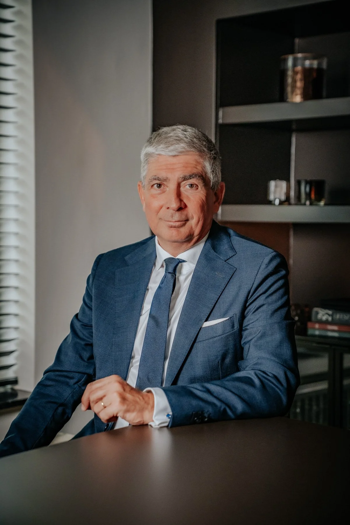 Een man in een wit overhemd staat met gekruiste armen in een interieur met een boog en houten elementen.