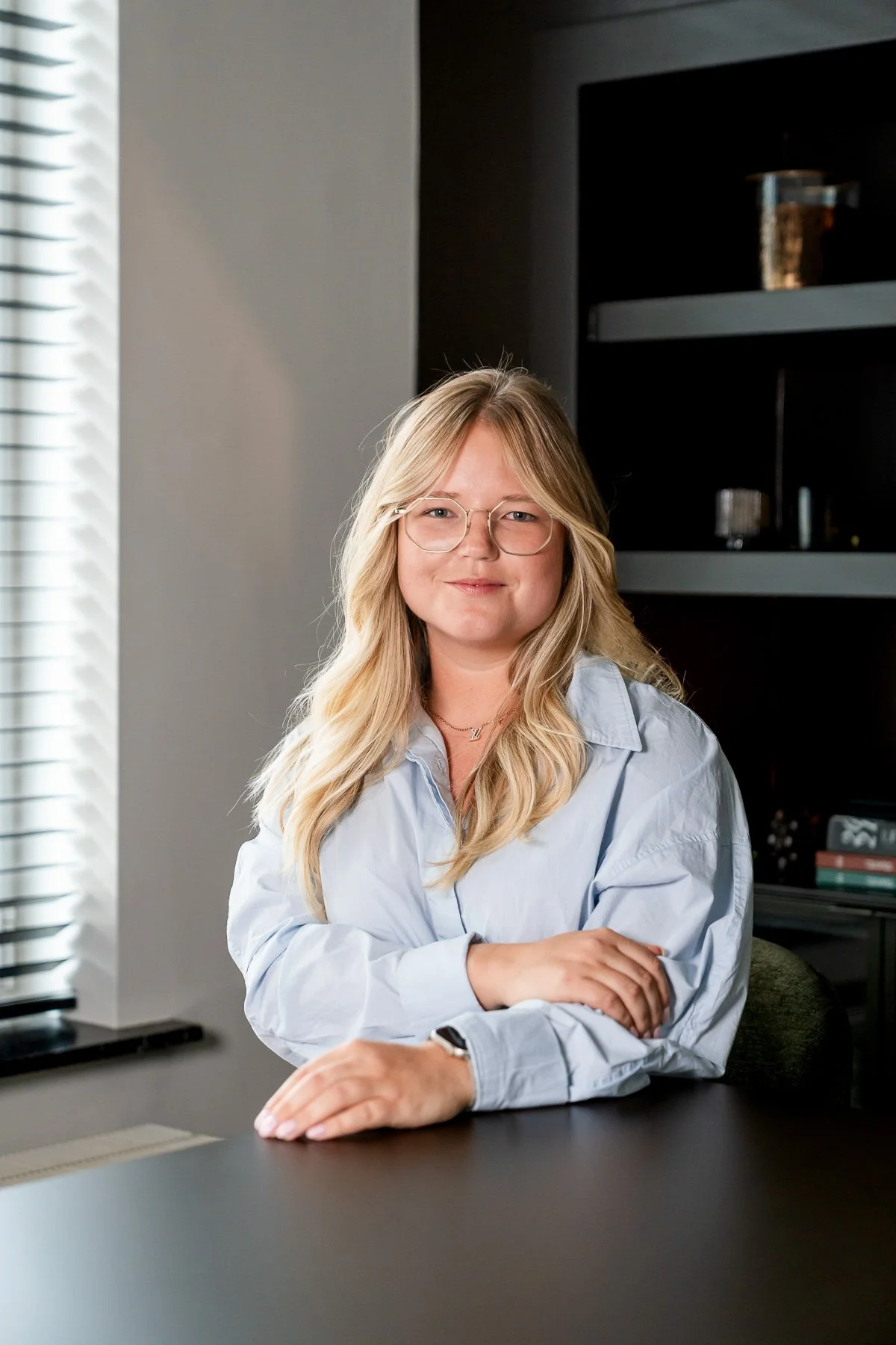 Een jonge vrouw met blond haar en brillen, gekleed in een zwarte jas, staat met haar armen gekruist in een kamer met een klassieke inrichting.