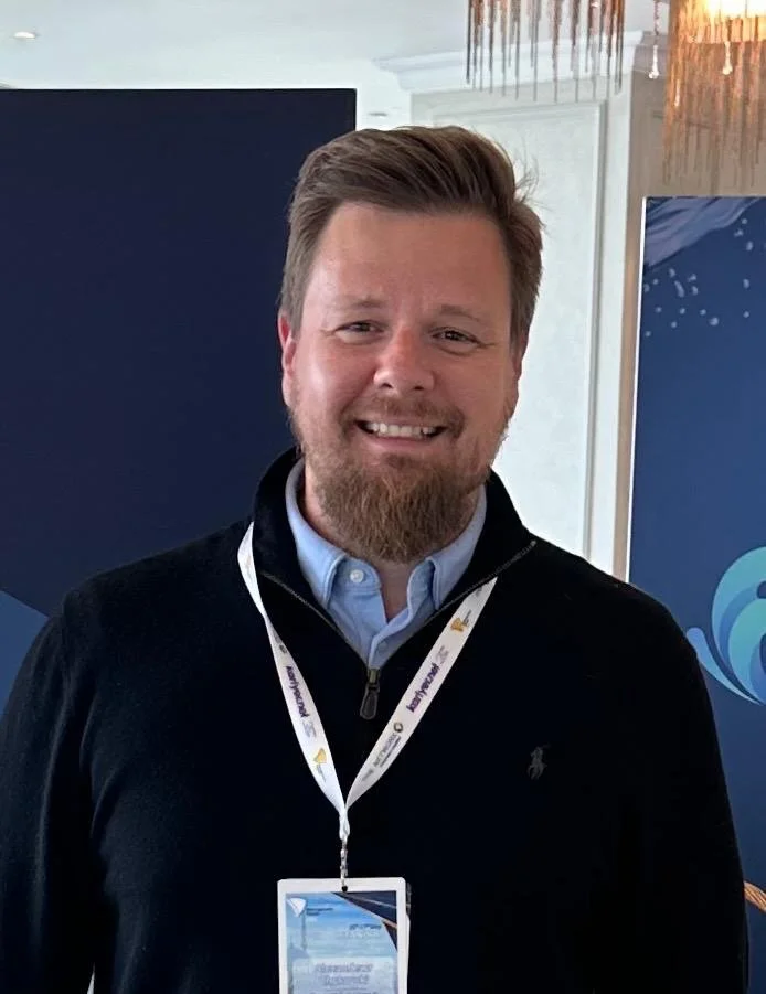 Smiling person with a beard wearing a black sweater and a white lanyard with a badge in an indoor setting.