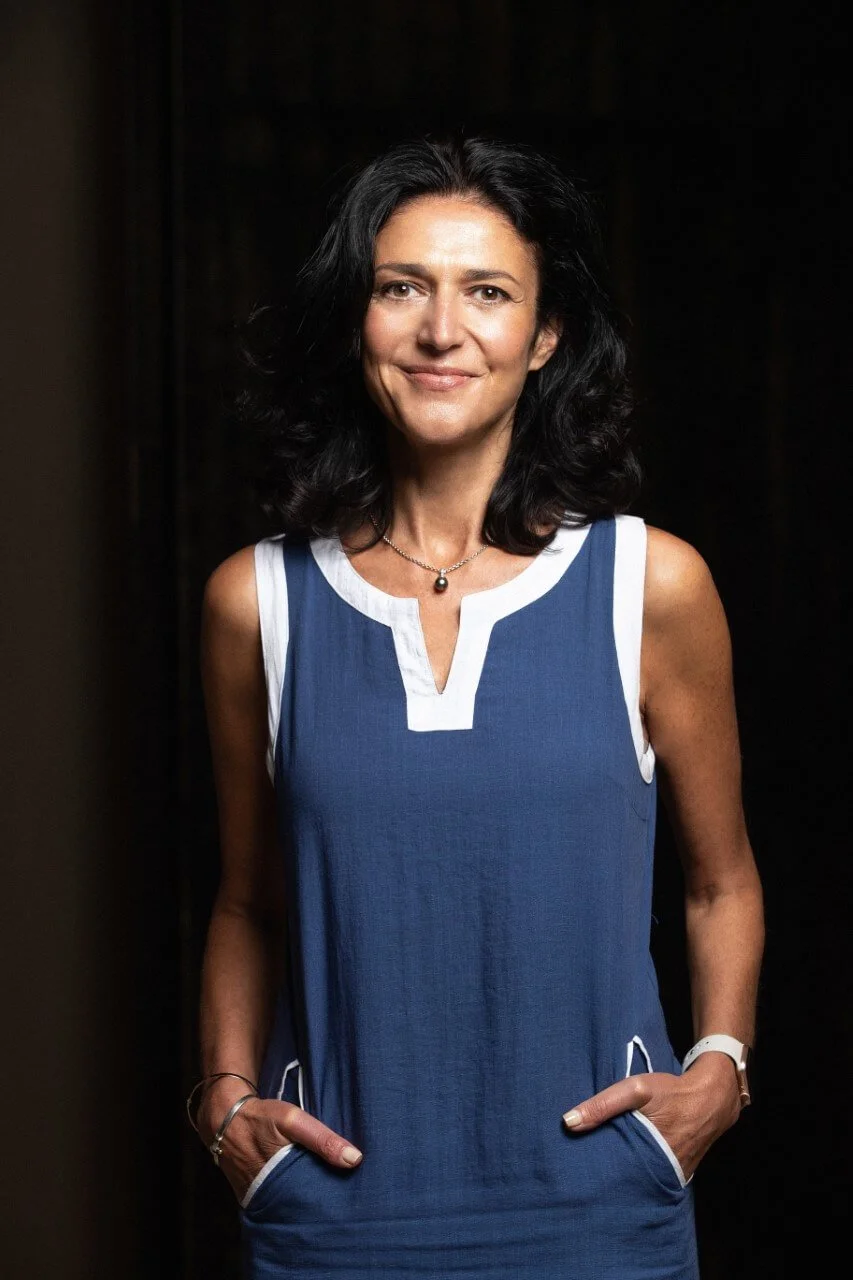 A woman with shoulder-length black hair and light skin, smiling and standing against a dark background. She is wearing a sleeveless blue dress with white trim and has her hands in her pockets. She is also wearing a necklace with a black pearl pendant and a white wristwatch.