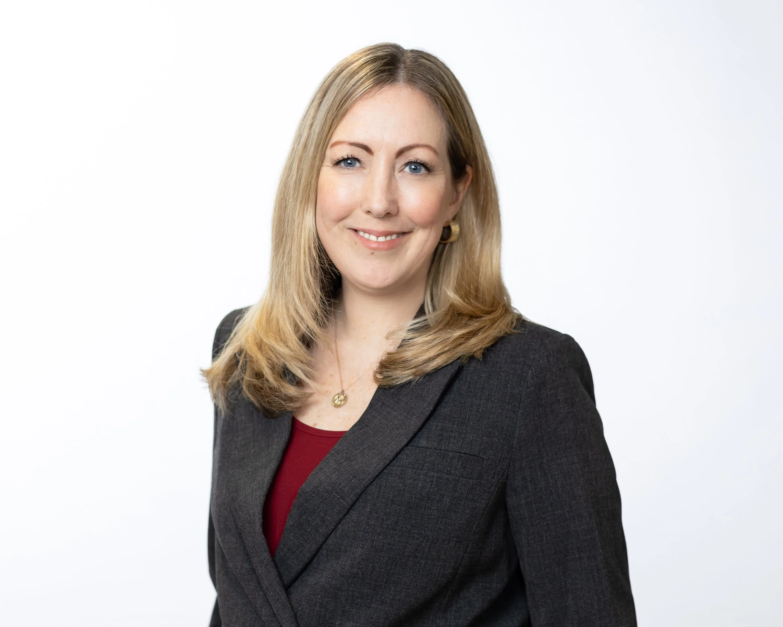 Professional woman with blonde hair wearing a dark blazer and red top, smiling against a plain white background.