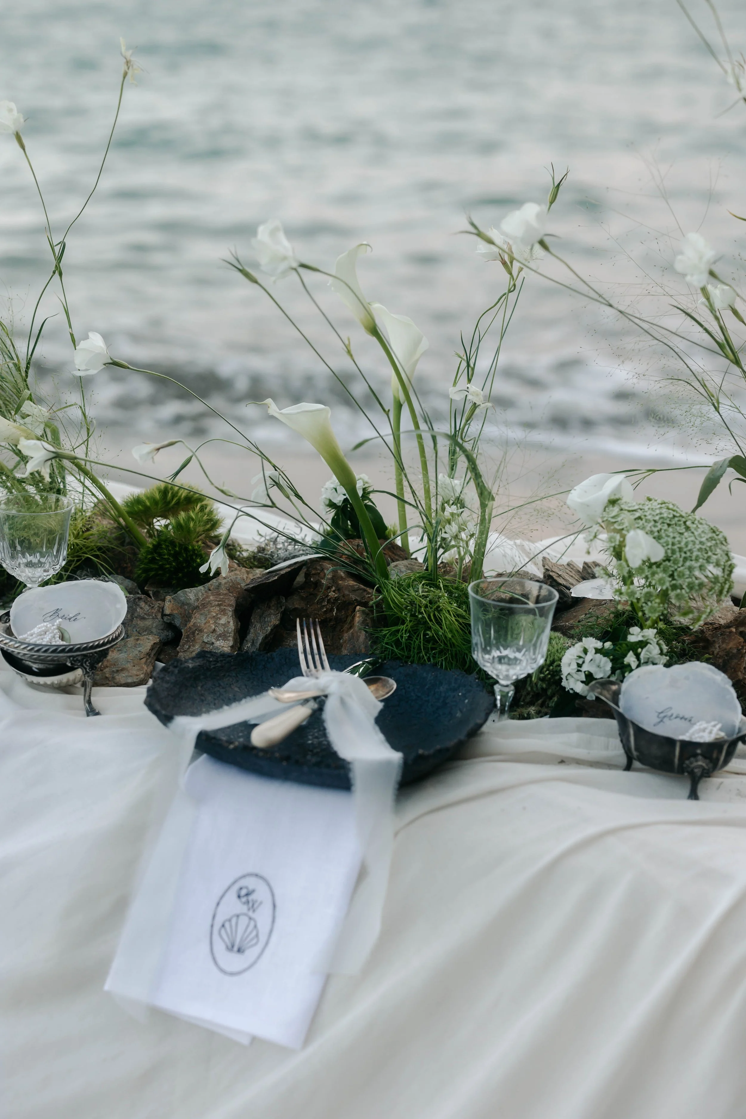 Elegant outdoor table setting with white flowers, glasses, and black plates on a cream tablecloth near the water.