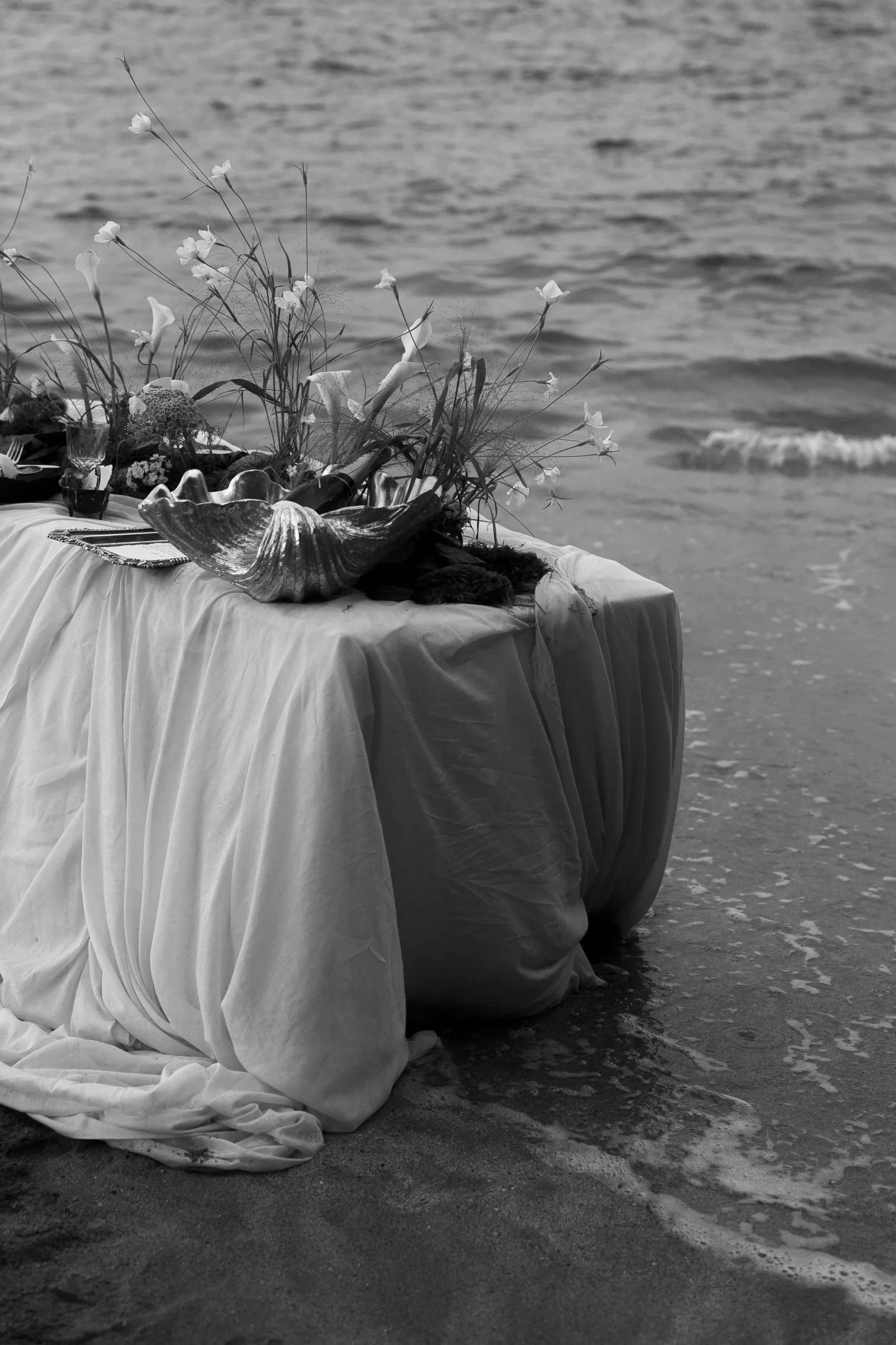 A table decorated with flowers, shells, and cloth on the beach near the ocean.