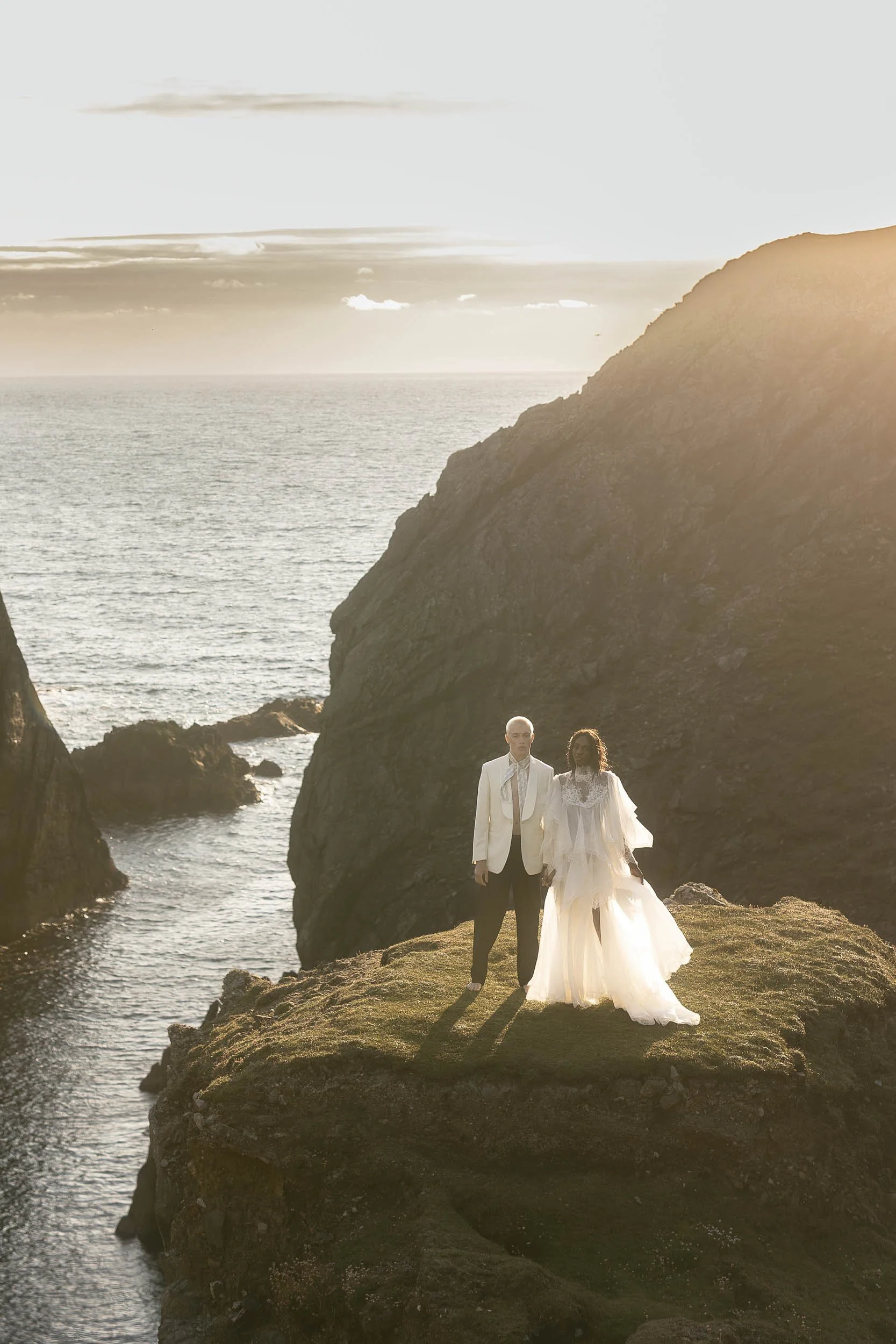 A couple dressed in wedding attire standing on a grassy rock formation with the ocean and cliffs in the background, illuminated by sunset.