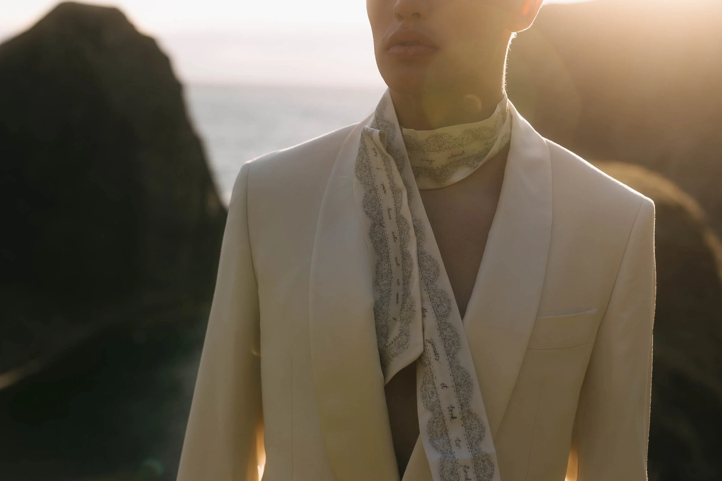 A person in a white blazer with a scarf around their neck standing outdoors during sunset, with rocks and the ocean in the background.