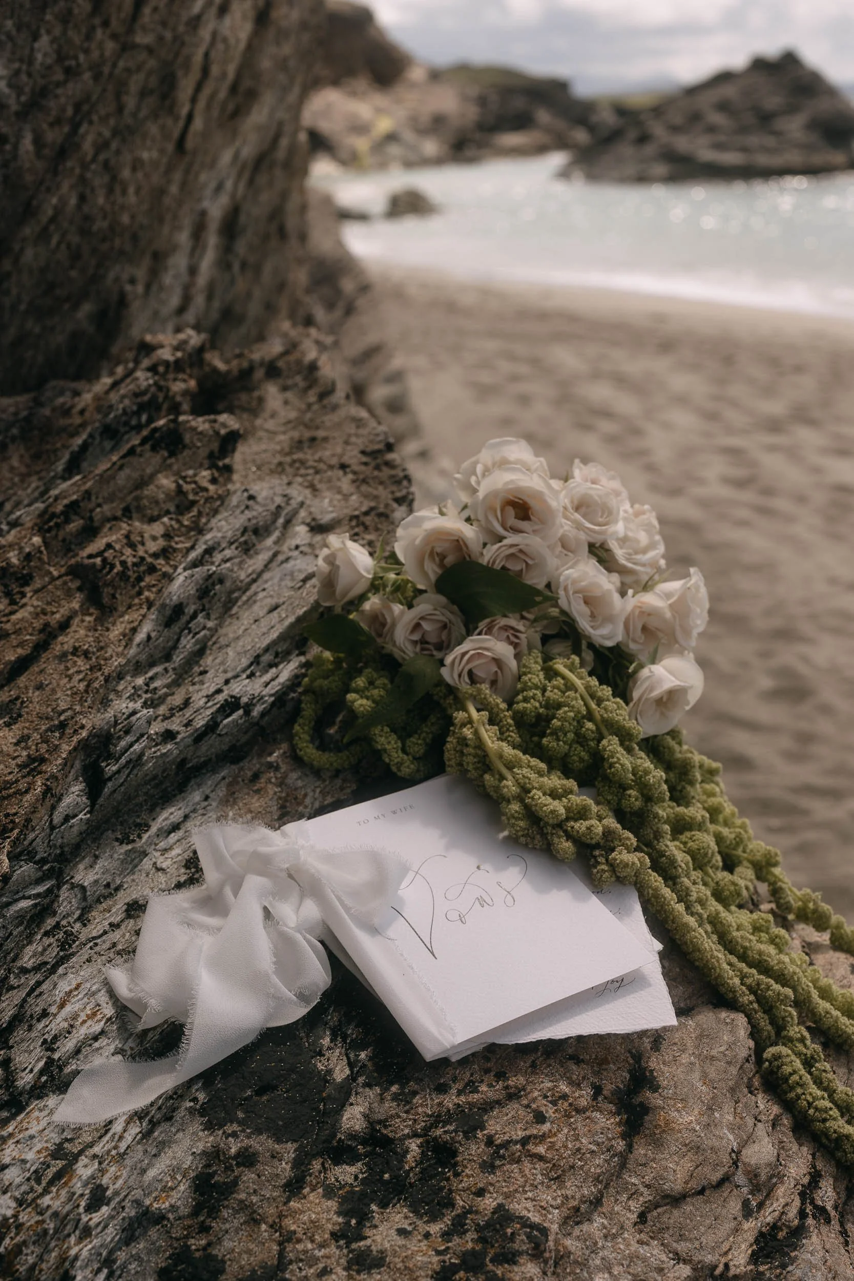 A bouquet of white roses with green foliage resting on a large, weathered rock near a beach with sandy shore, rocks, and ocean in the background.