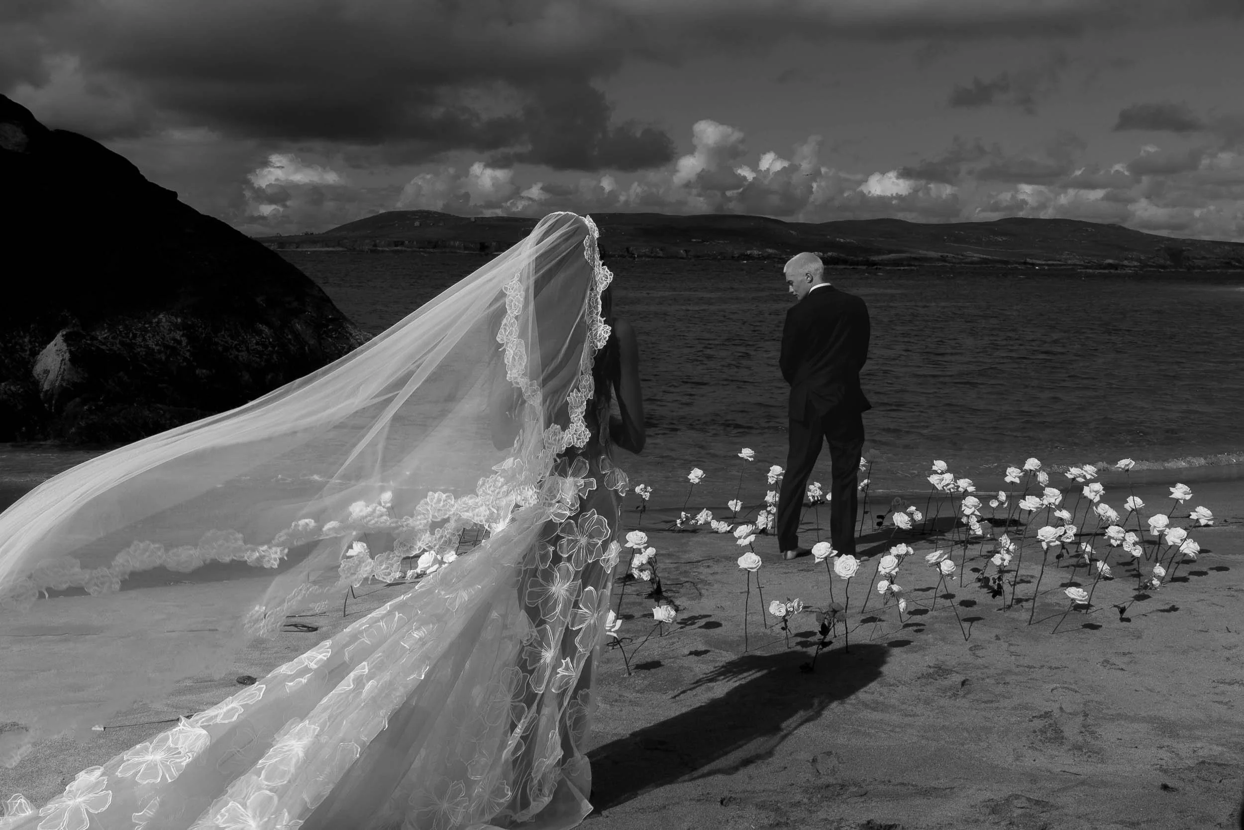 A bride in a wedding gown with a long veil and a groom in a suit standing on a beach with flowers, rocks, and water, under a cloudy sky.