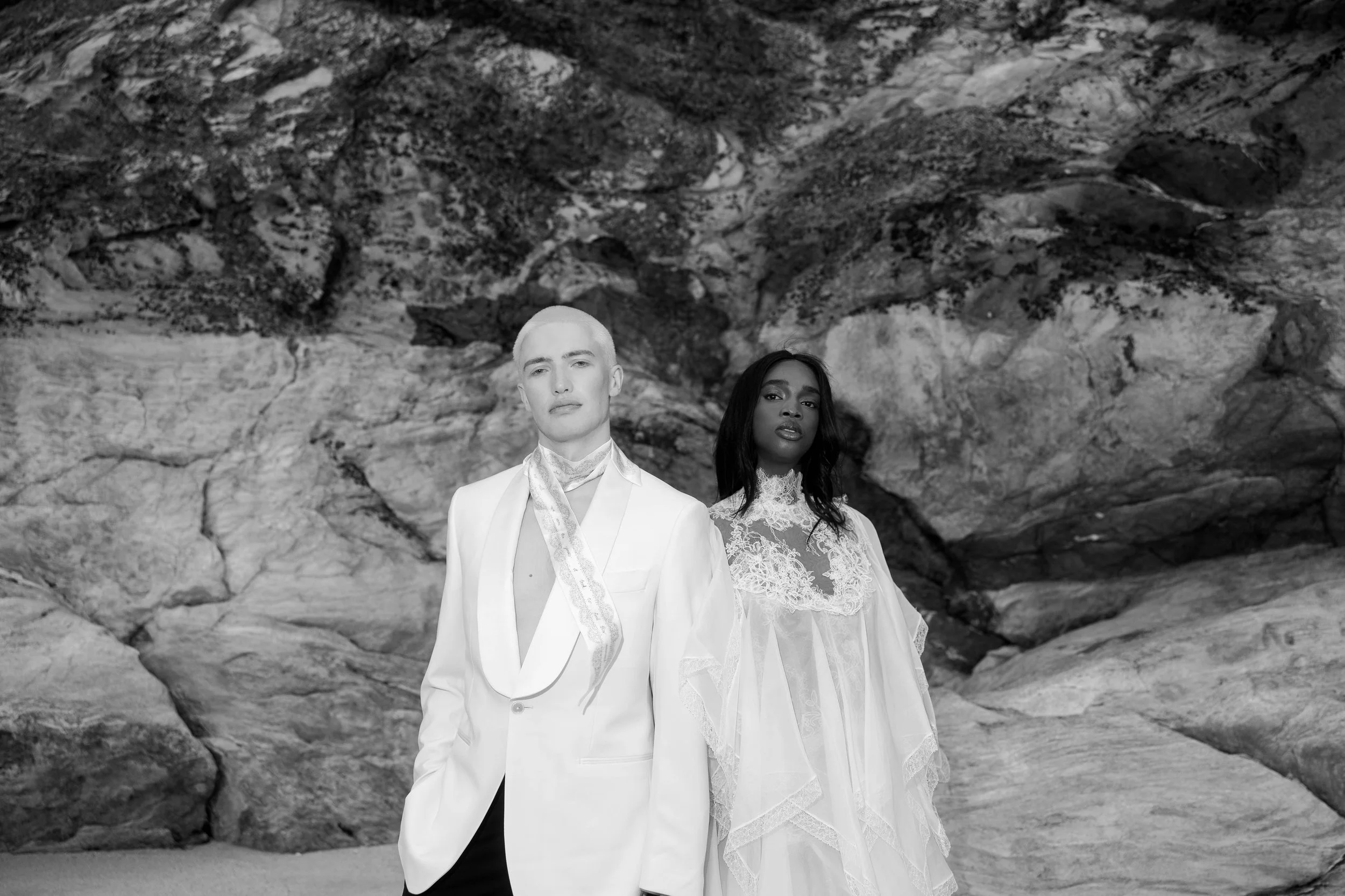 Black and white photograph of a man and woman standing in front of a rocky background, dressed in formal attire. The man wears a white suit with an open collar and a patterned scarf, while the woman wears a lace dress.