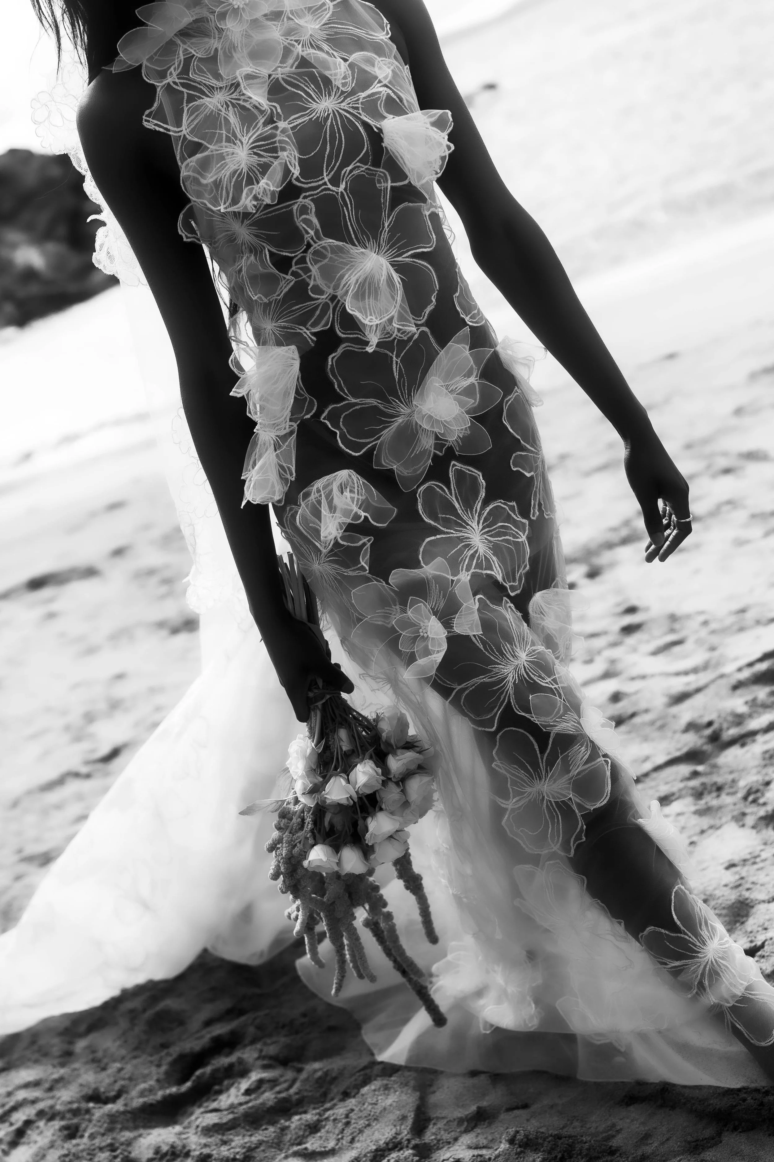 A woman in a sheer, floral-embellished dress holding a bouquet of flowers, standing on a beach with sand and ocean in the background.