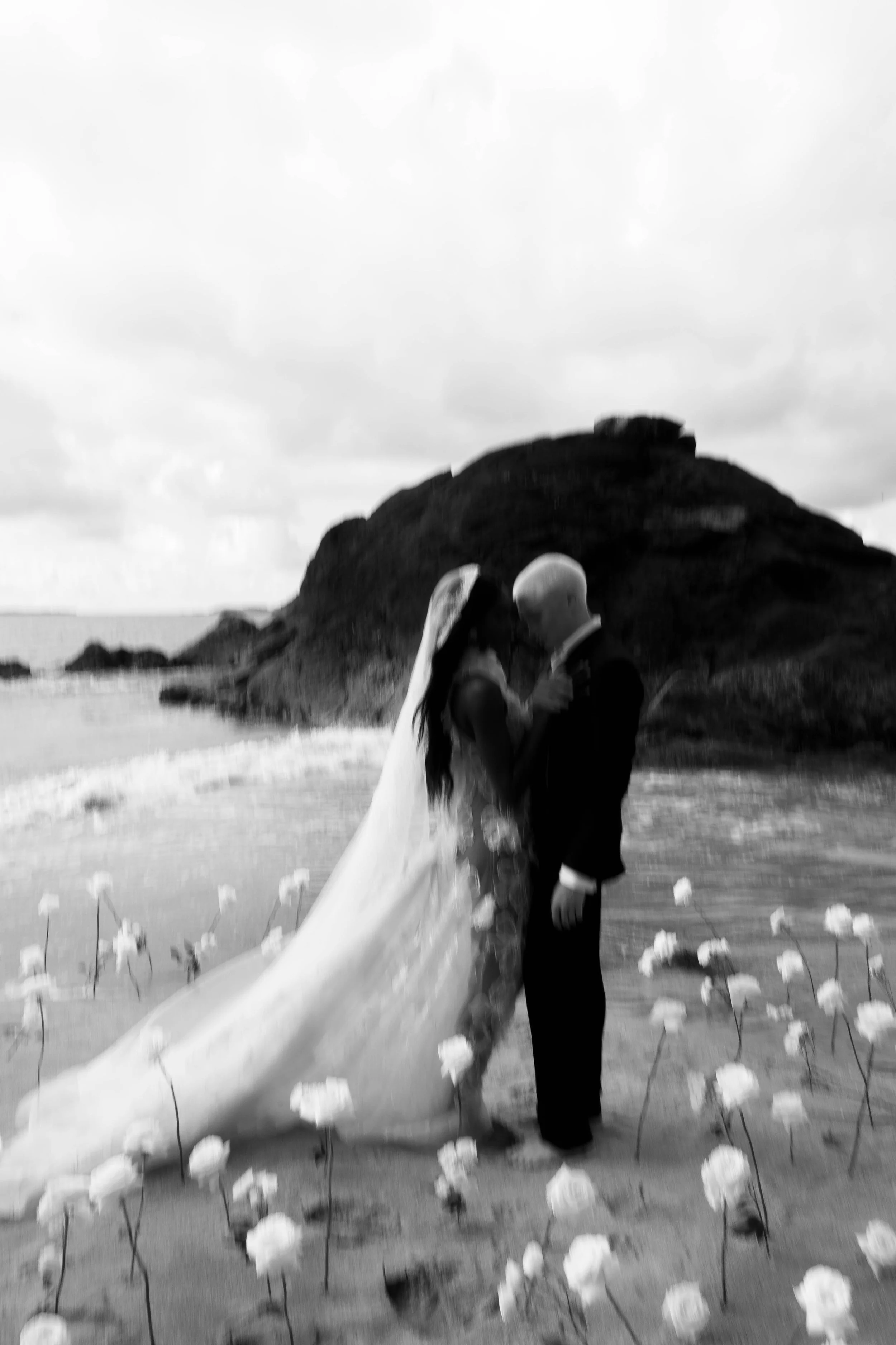 Black and white photo of a bride and groom standing in shallow water on a beach with a rocky hill in the background, holding hands and gazing at each other.