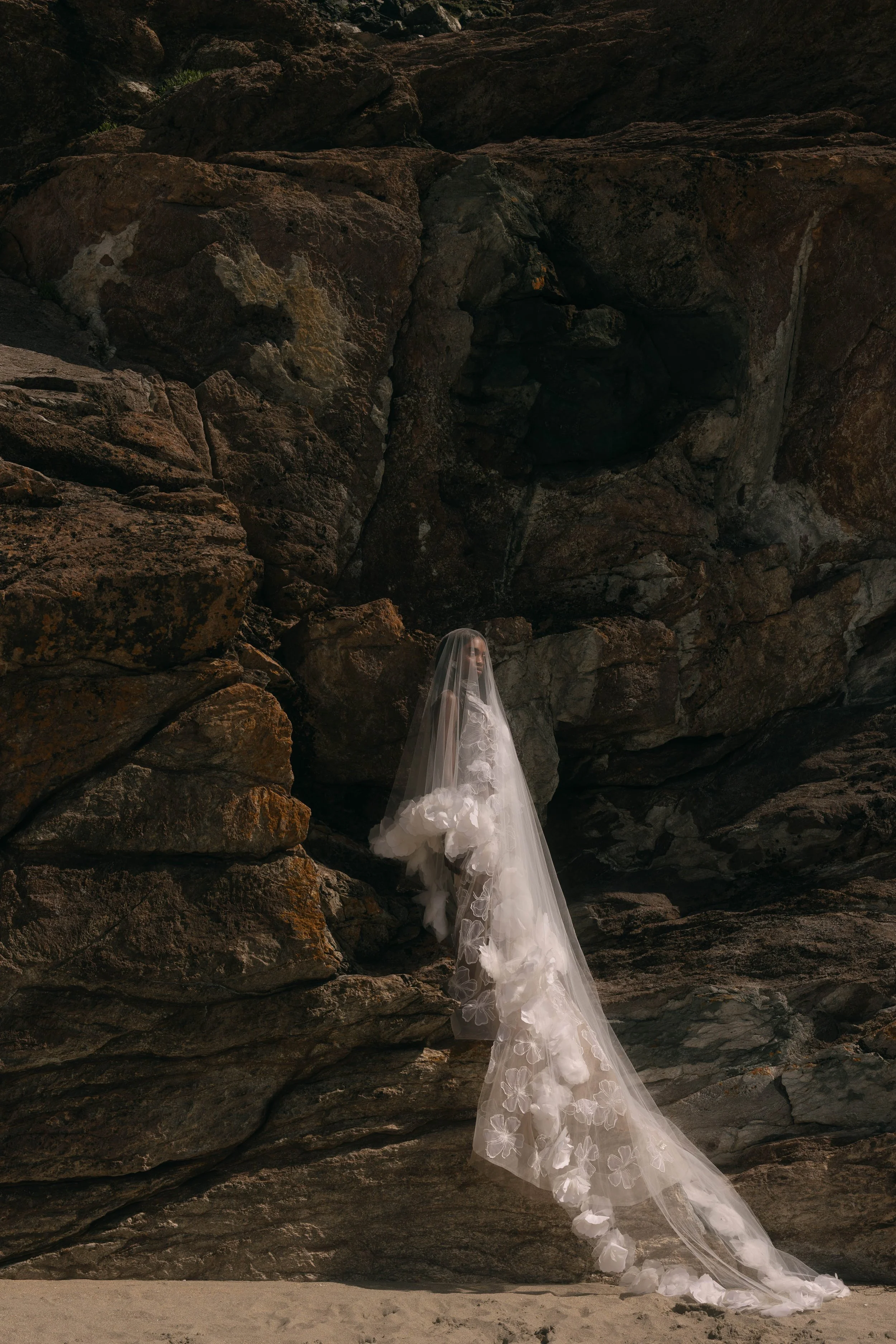A woman in a wedding dress and veil stands on a rocky beach near a large cave or rock formation.