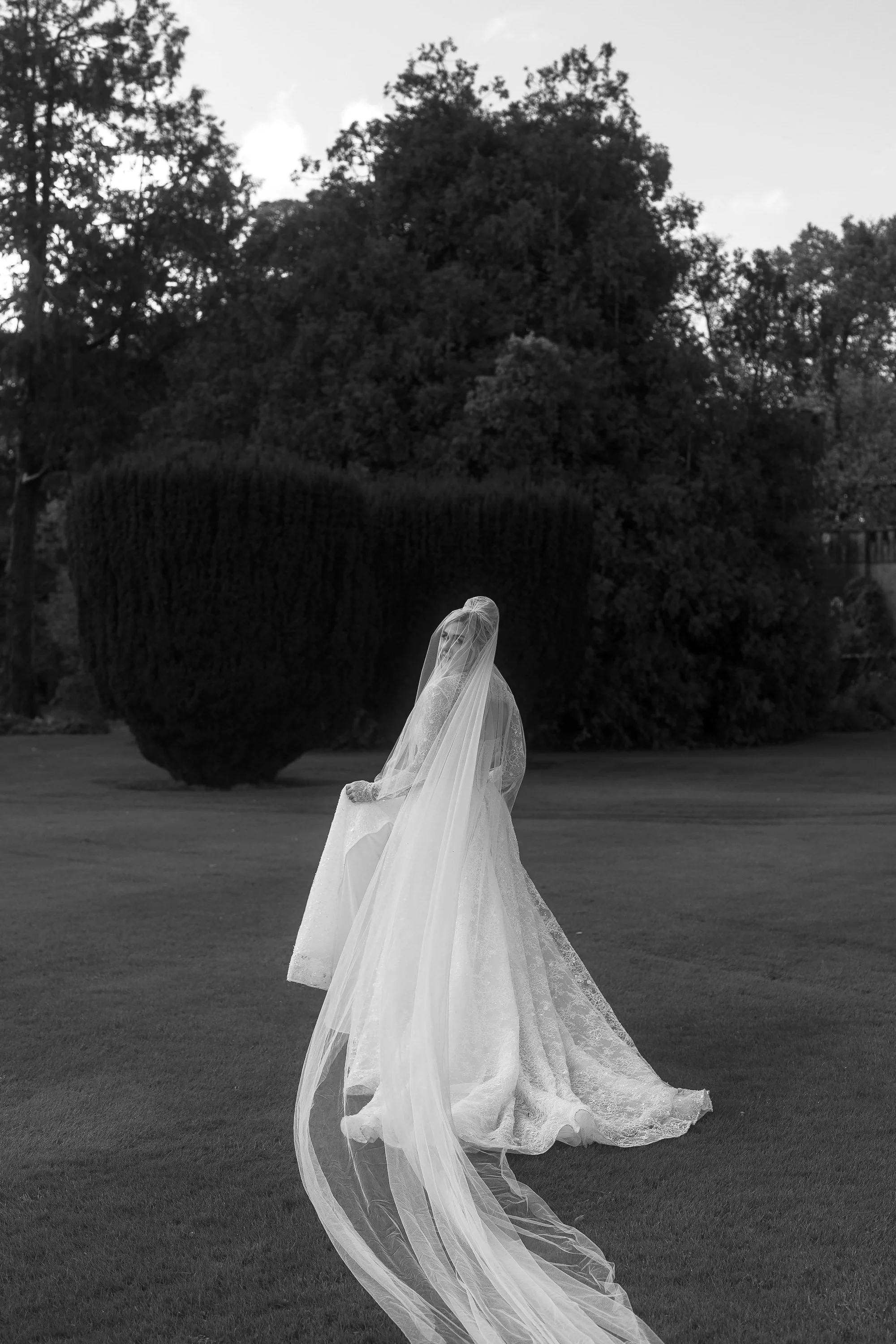 A bride in a wedding dress and veil standing on a grassy lawn with trees and bushes in the background.