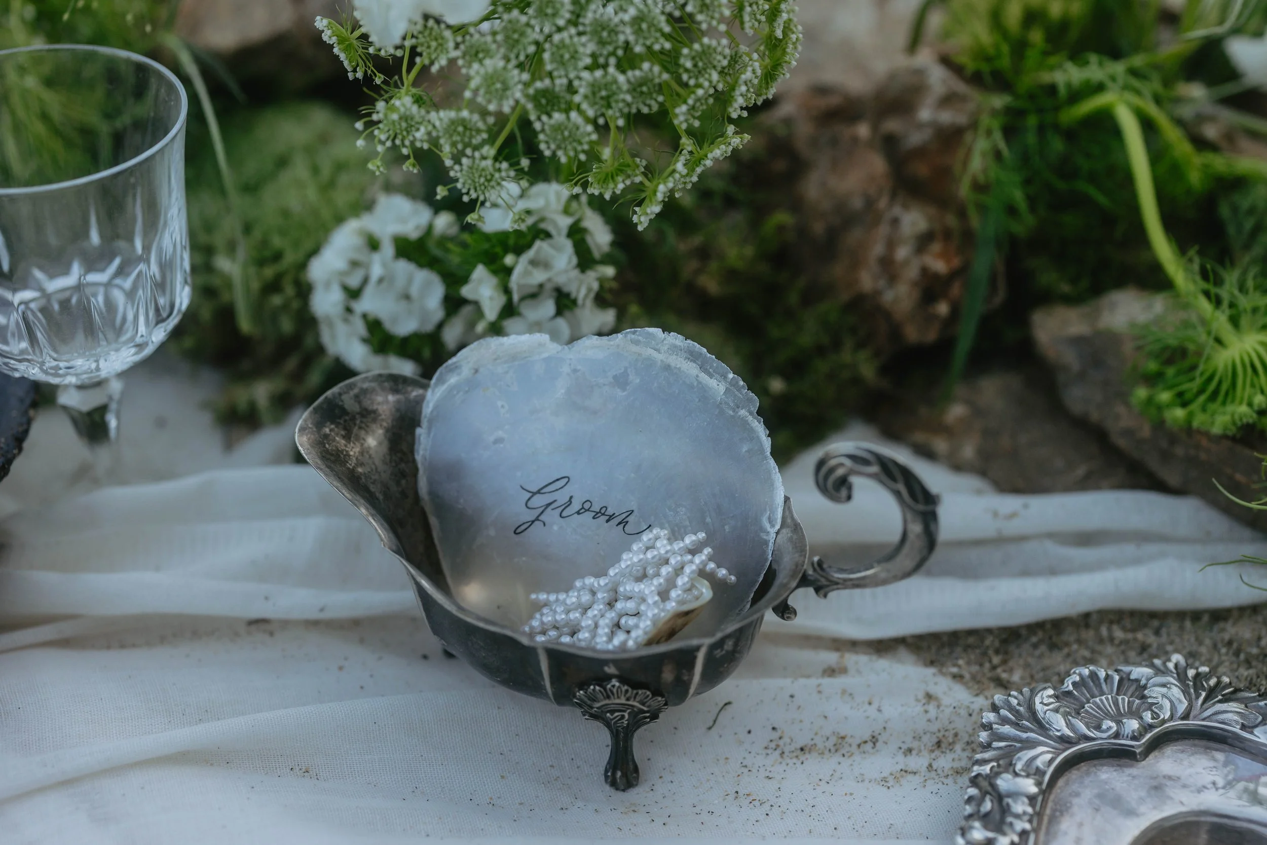 A silver dish labeled "Groom" holding pearl necklaces on a white cloth, decorated with flowers and greenery, at an outdoor wedding setup.