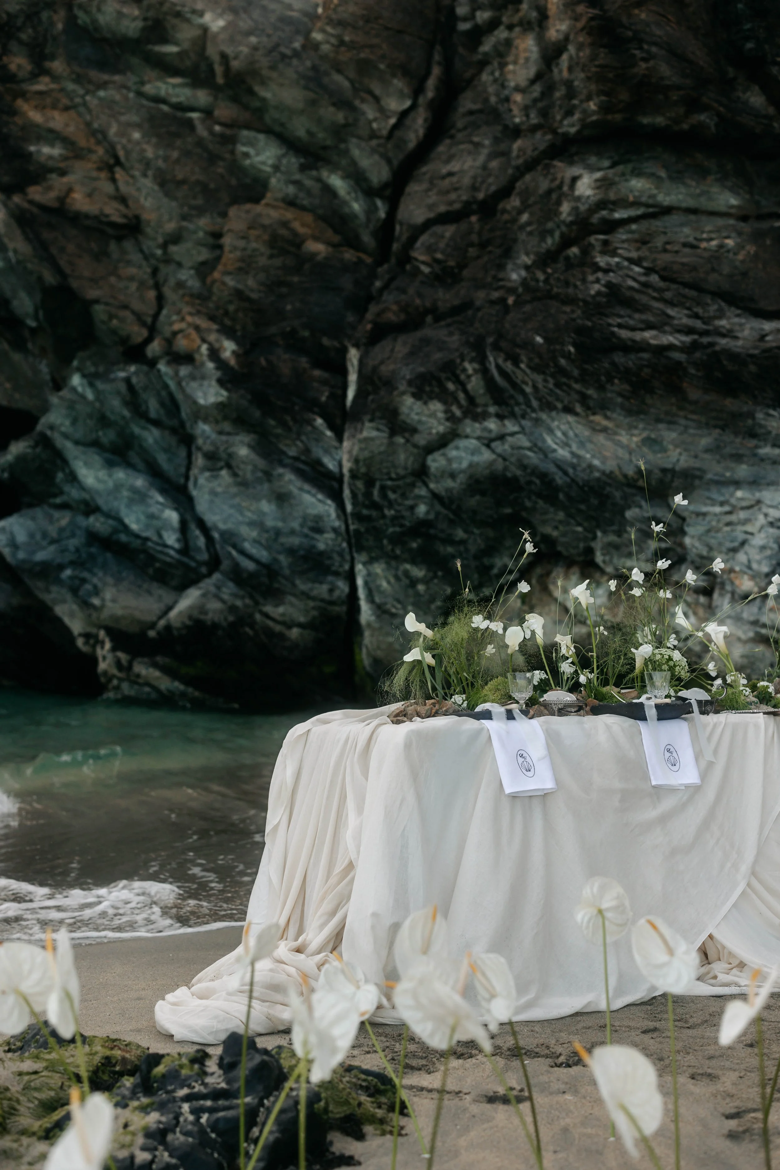 A beachside wedding altar with white flowers, glassware, and embroidered cloths set against a rocky cliff background.