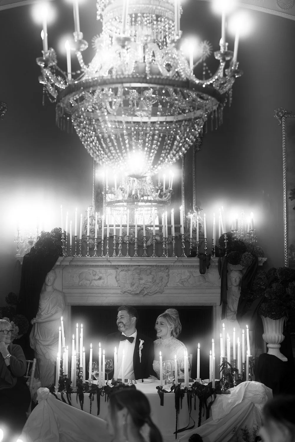 A wedding reception with a bride and groom sitting at the head table, surrounded by tall candles and elegant decorations, in a grand hall with a large chandelier overhead.