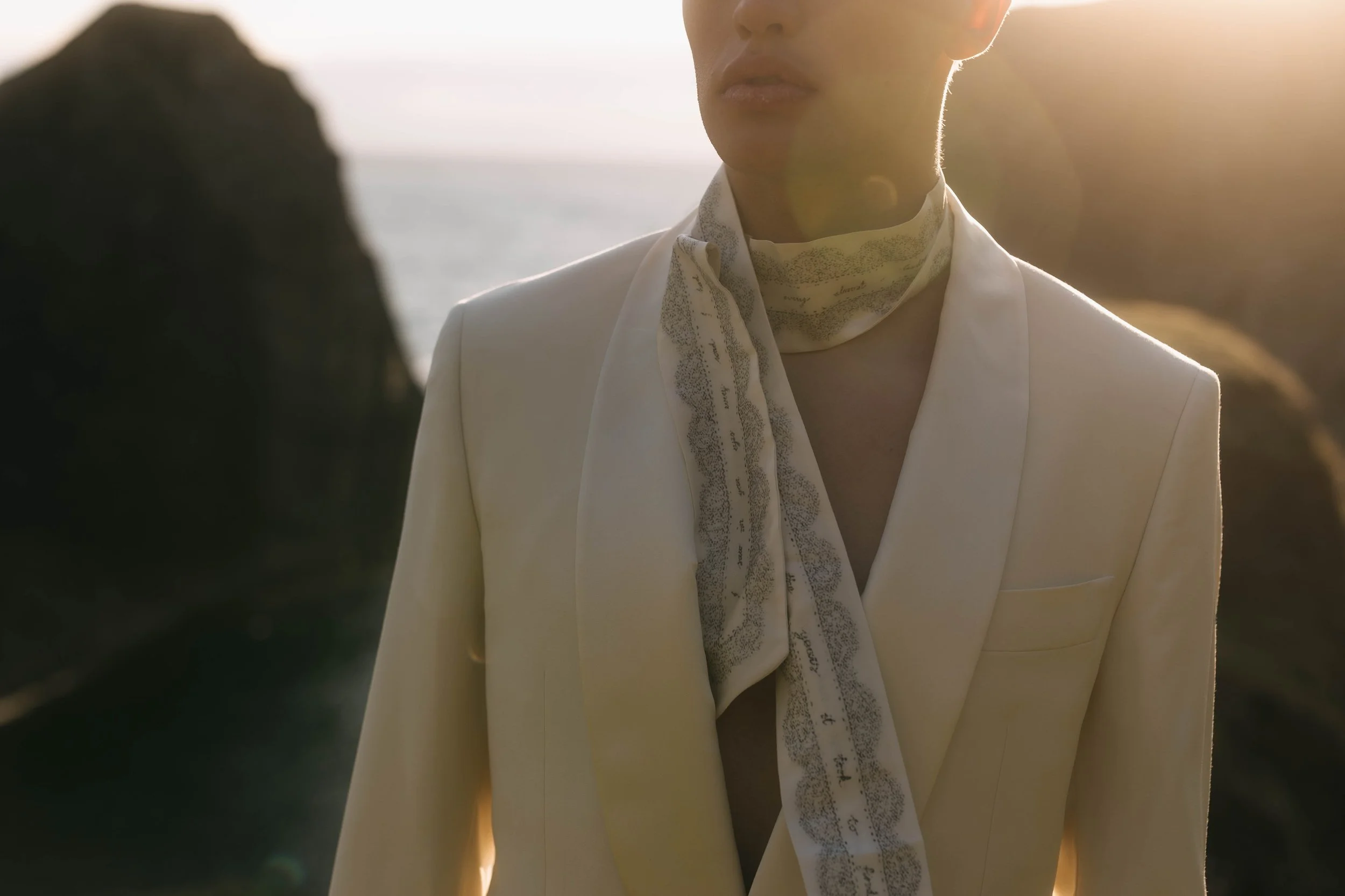 Person wearing a white blazer and a patterned scarf standing outdoors near rocky cliffs and water with sunlight