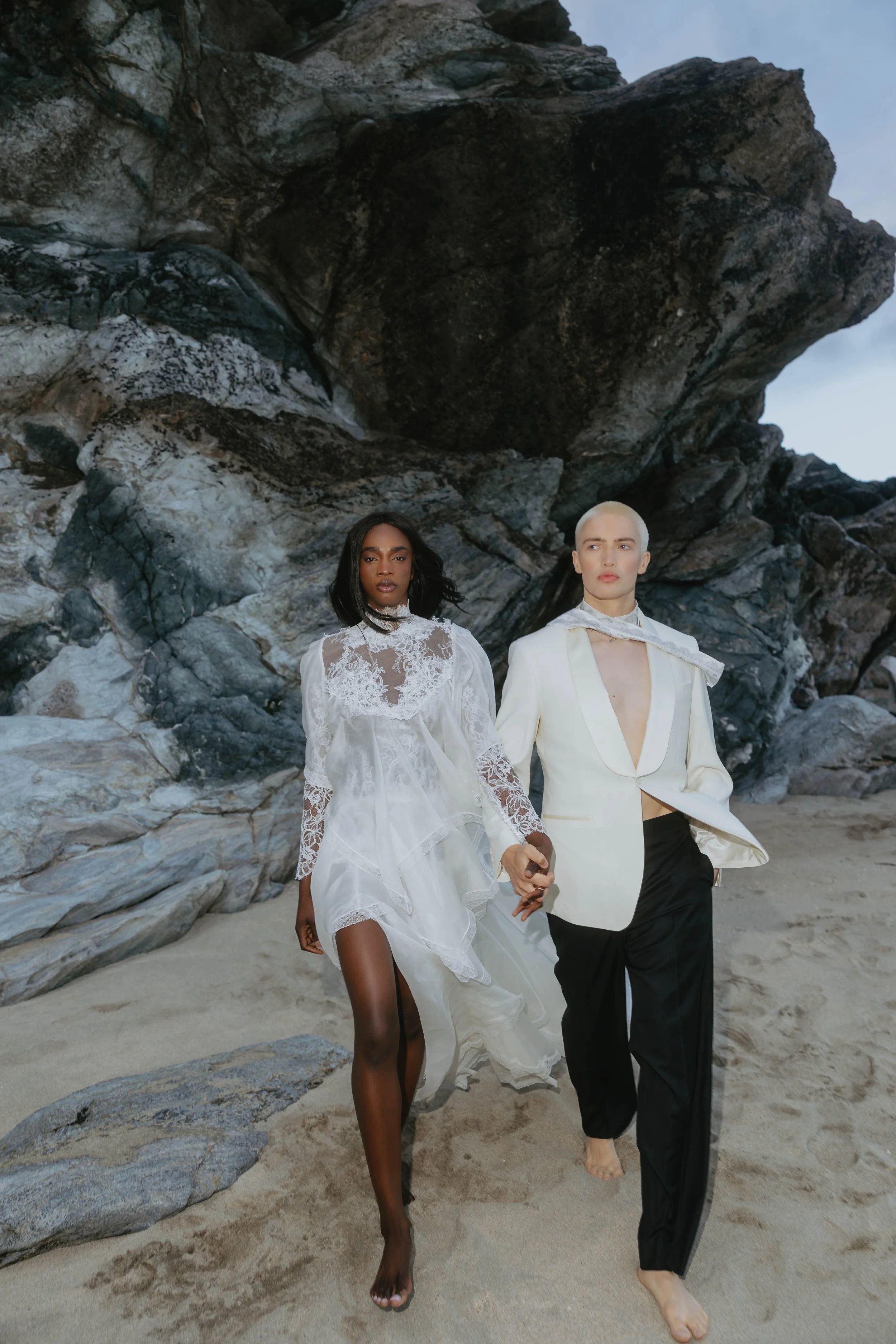 Two models, a woman and a man, walking hand in hand on a sandy beach with large rocks and a cliff in the background. The woman is wearing a white lace dress, and the man is dressed in a white blazer and black pants.