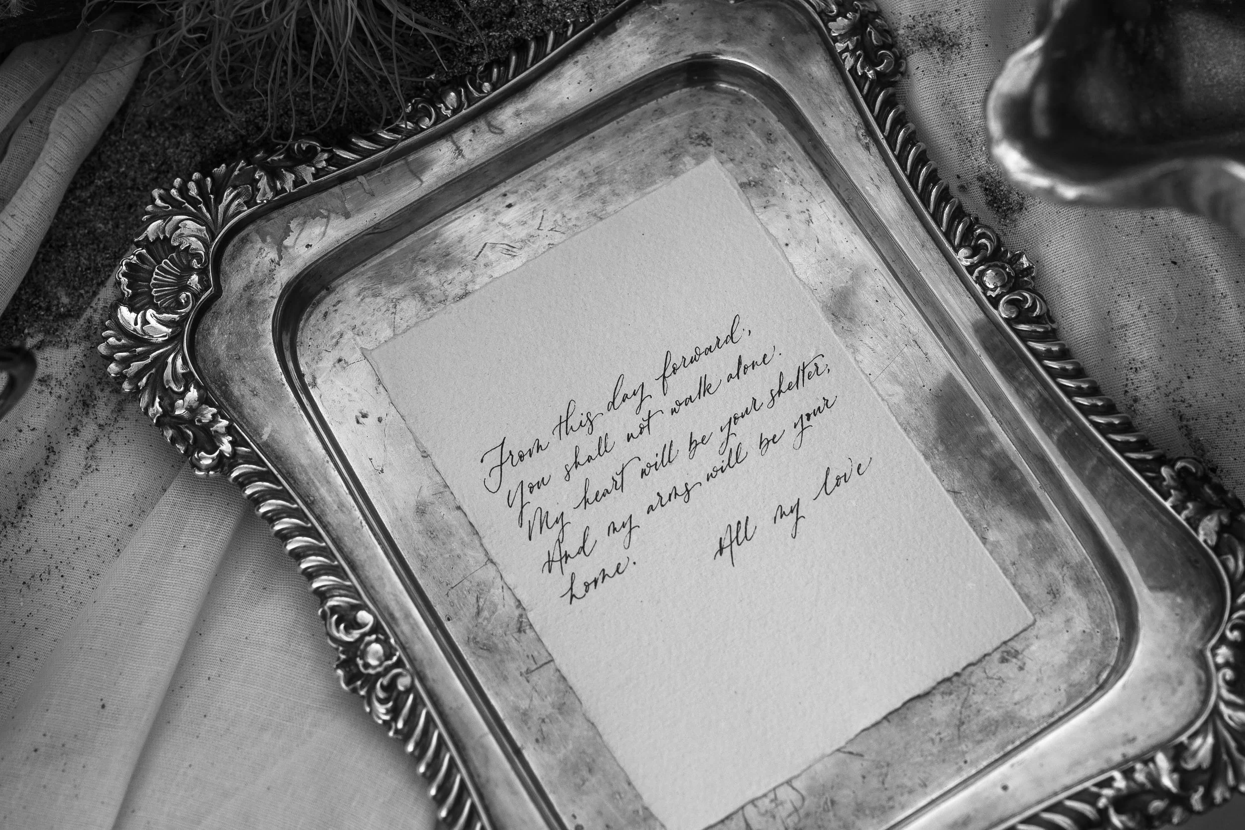A silver decorative tray with an ornate border holding a handwritten note on a piece of paper.