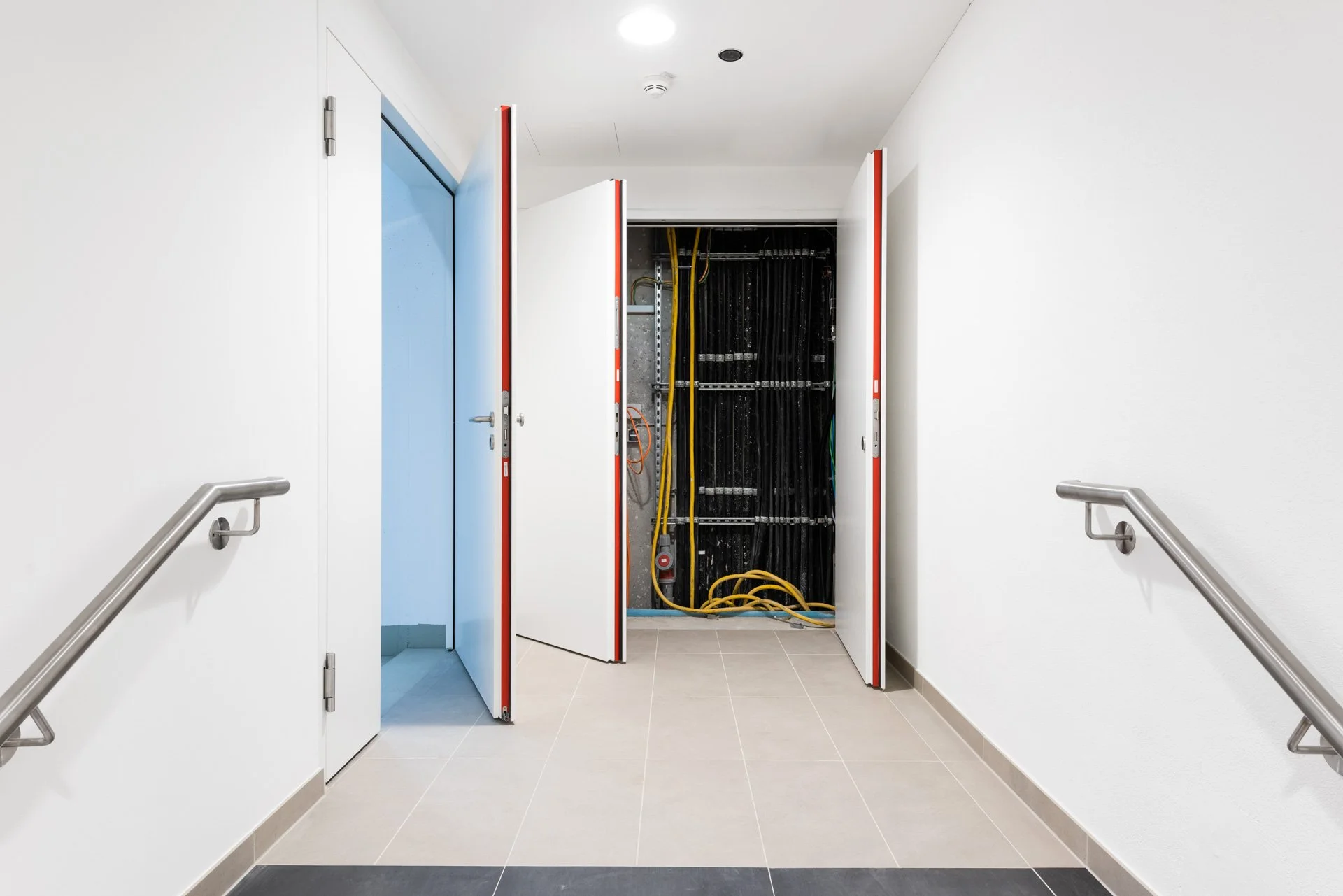 Ein Flur mit weißen Wänden und einem schwarzen Boden. Zwei weiße Türen, eine auf der linken Seite und eine offene Tür in der Mitte, führen zu einem Technikraum mit schwarzen Kabeln und gelben Kabeln an der Wand.