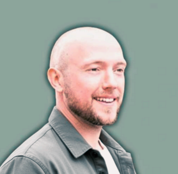 A smiling man with a beard and no hair, wearing a gray jacket, standing against a plain background.