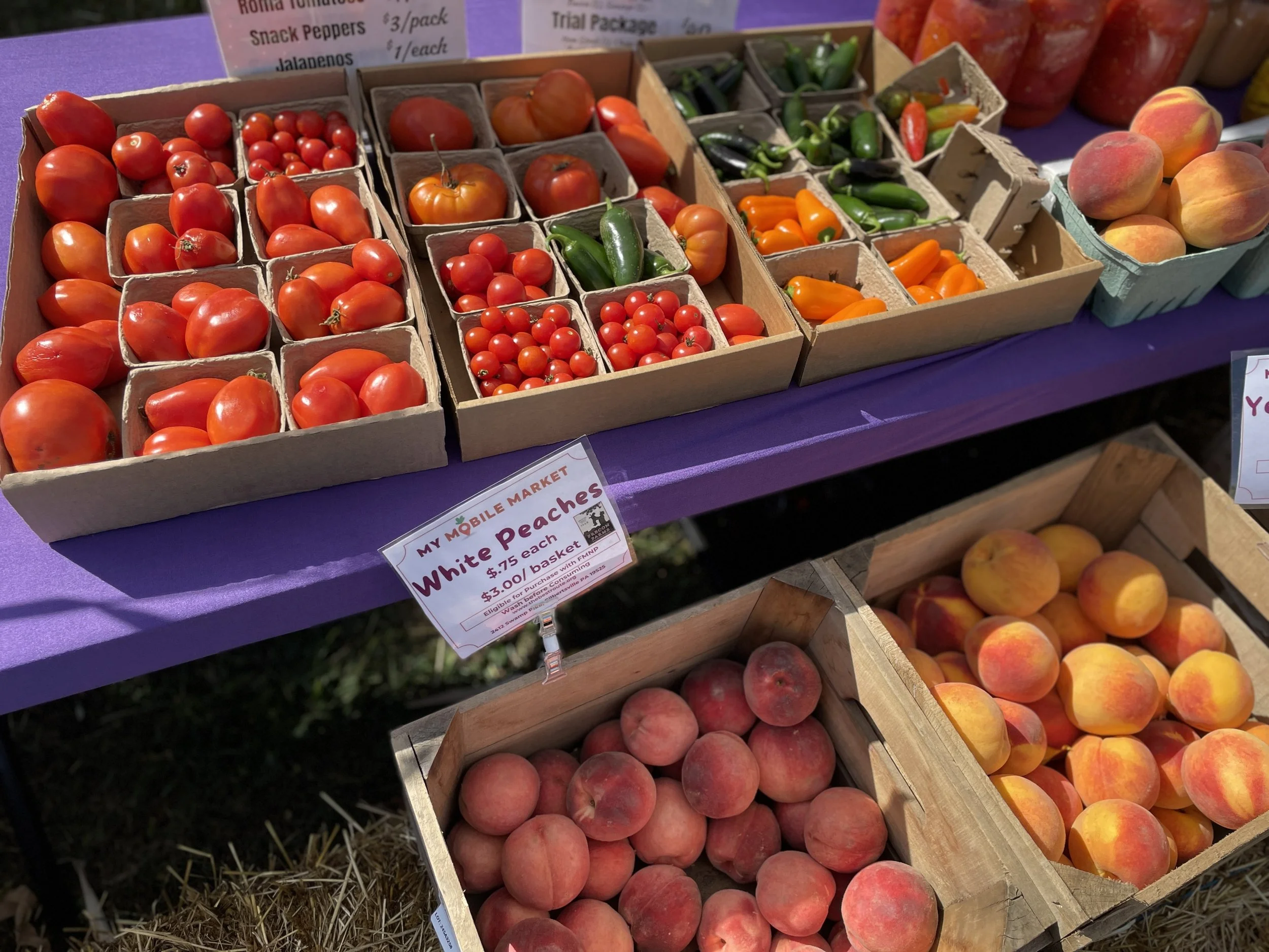 Boxes of fresh tomatoes, including cherry, grape, and larger varieties, along with peaches displayed on a purple table at a farmers market