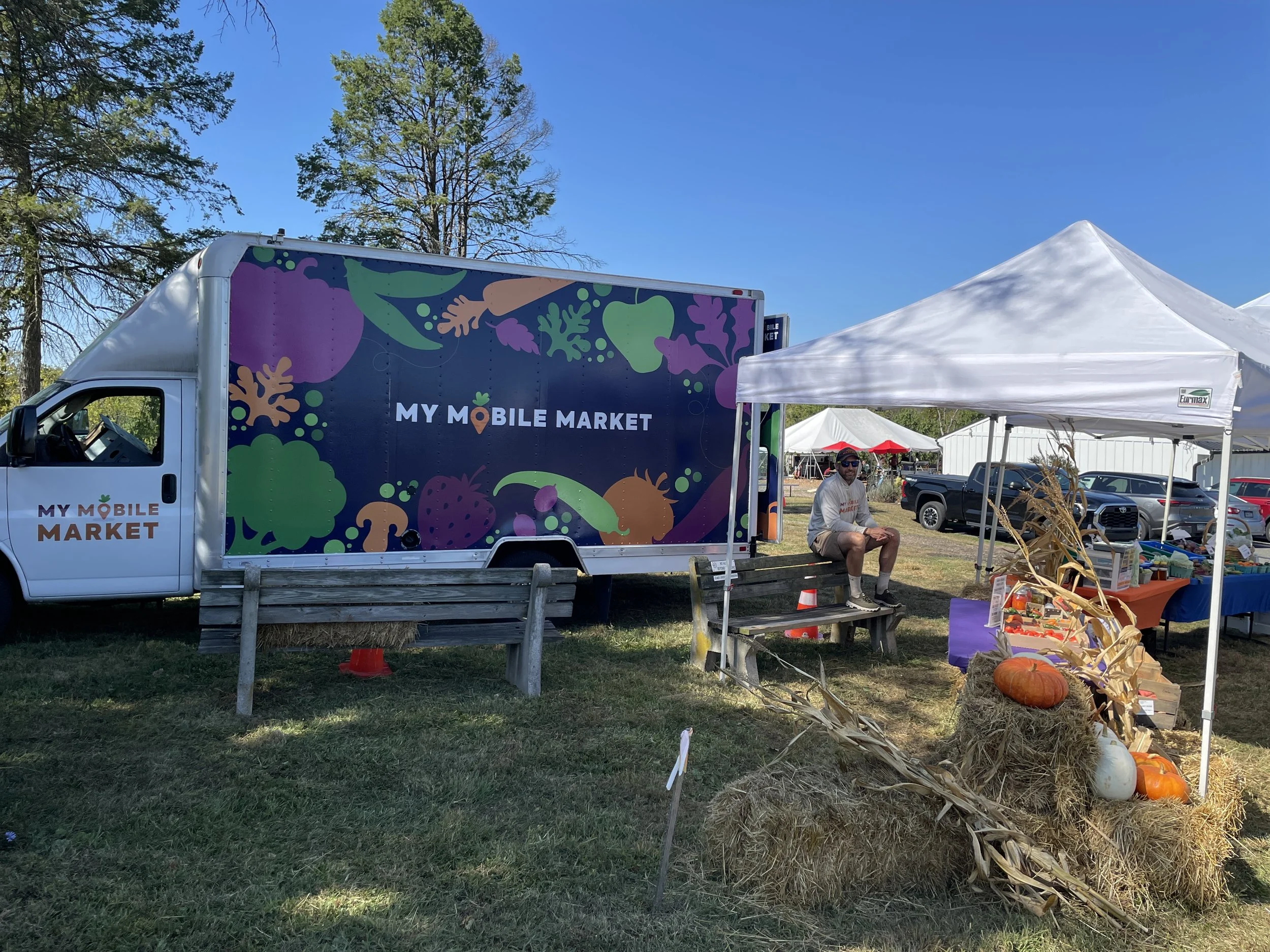 A mobile market truck with colorful fruit and vegetable illustrations and the text "My Mobile Market" parked at an outdoor market. There is a small white tent with a man sitting on a bench, and a display with pumpkins, hay bales, and dried corn stalks.