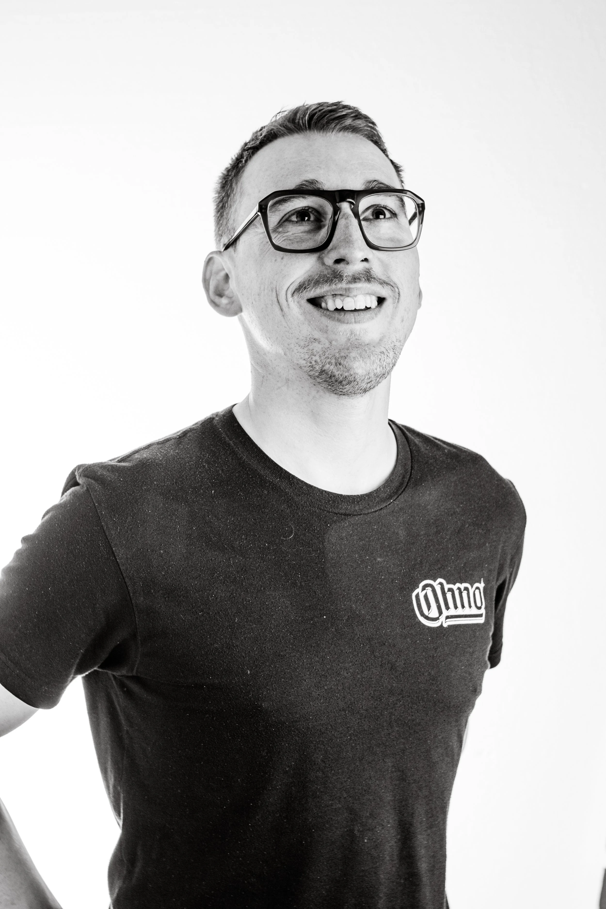 Black and white photo of smiling young man wearing glasses and a black t-shirt with a logo on the chest.